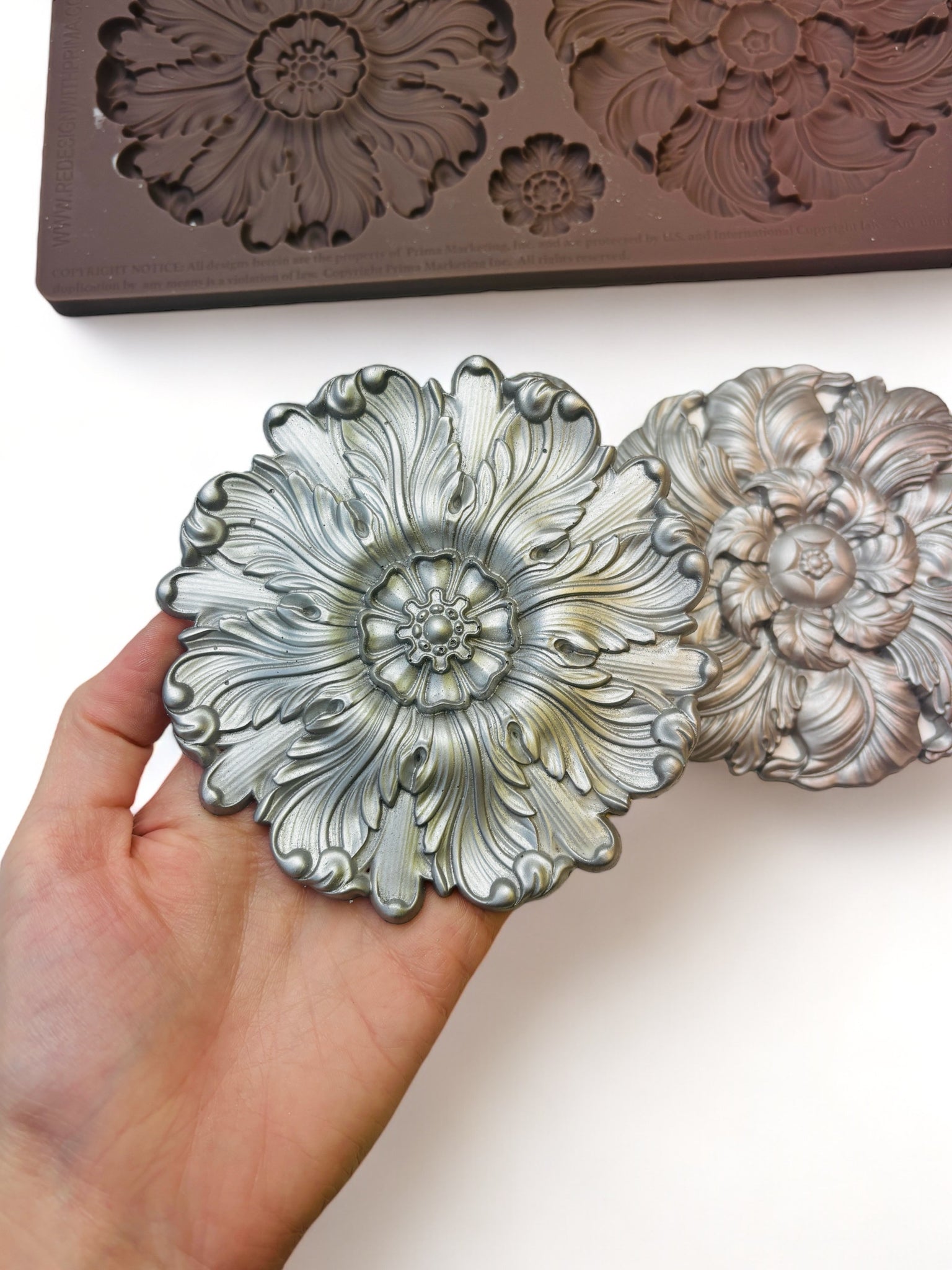 A brown silicone mold and silver colored castings of 2 large and 2 small floral medallions are against a white background. A hand is shown holding one of the large medallions.