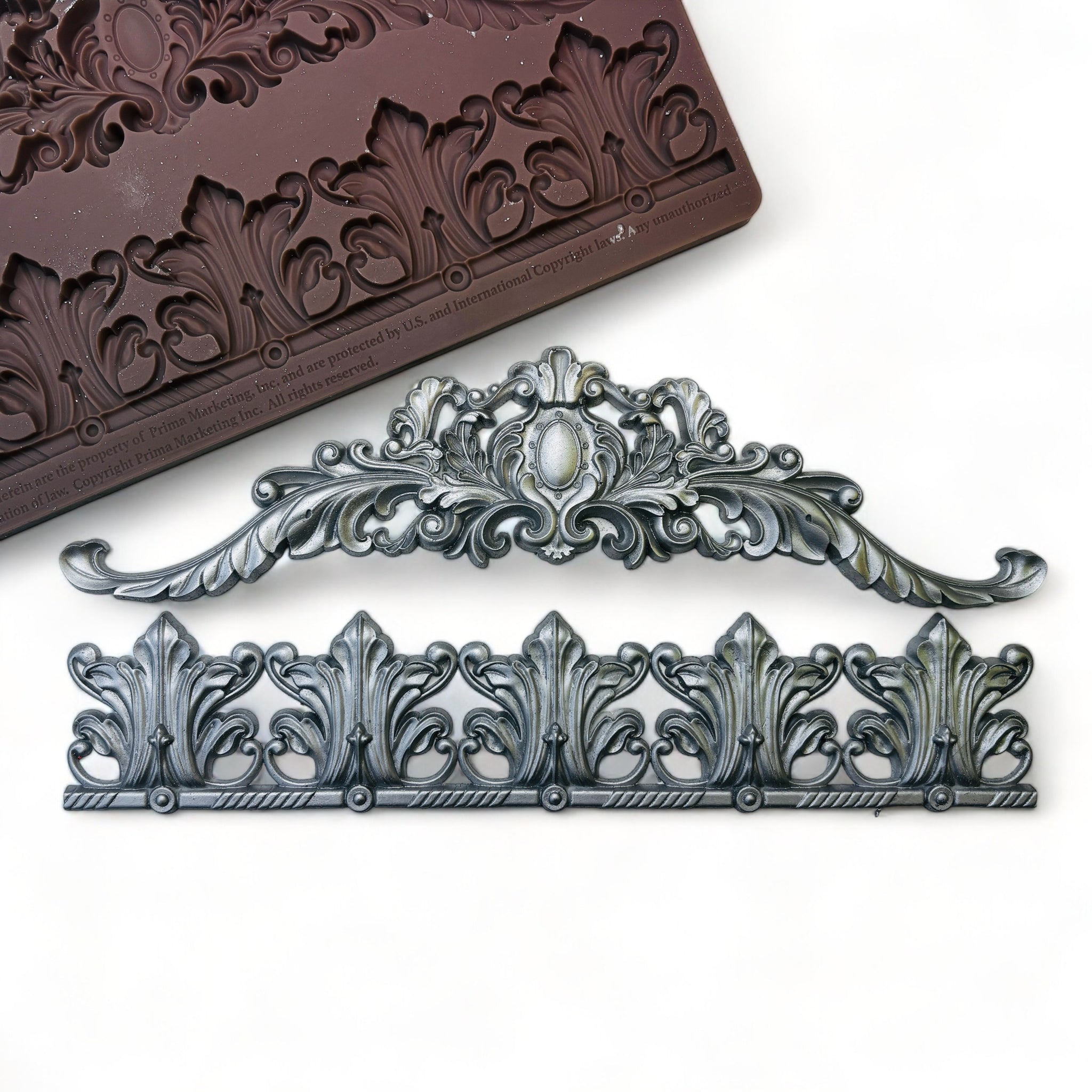 A brown silicone mold and silver colored casting of an ornate flourish repeating border trim and an ornate feather scroll center piece are against a white background. 