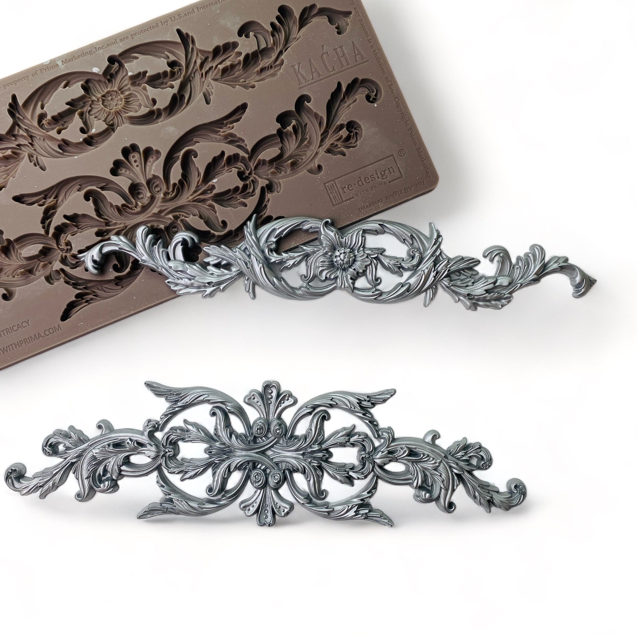 A brown silicone mold and silver colored castings of 2 ornate scrolls are against a white background.