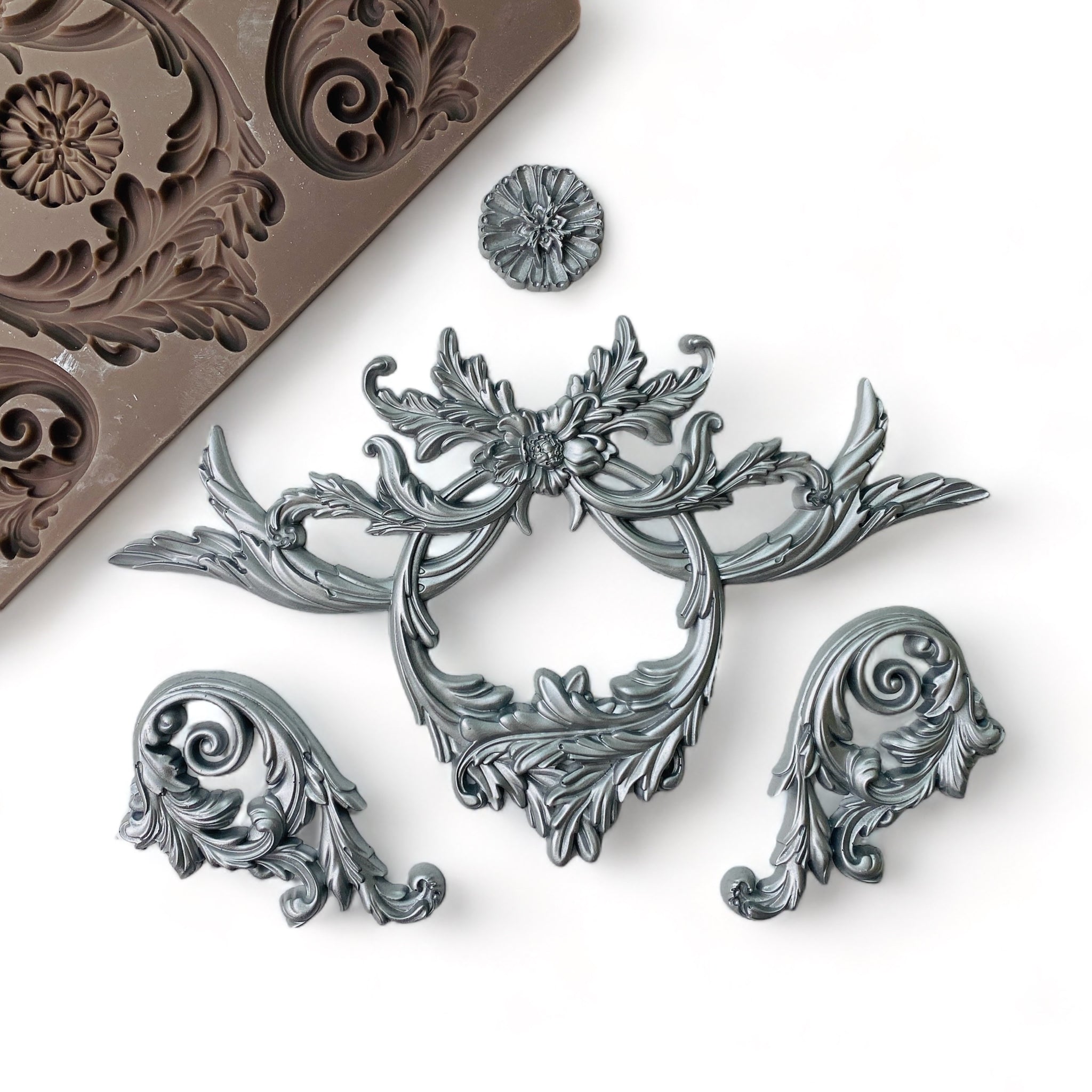 A brown silicone mold and silver colored castings of ornate scrolling accent pieces and a medallion are against a white background.