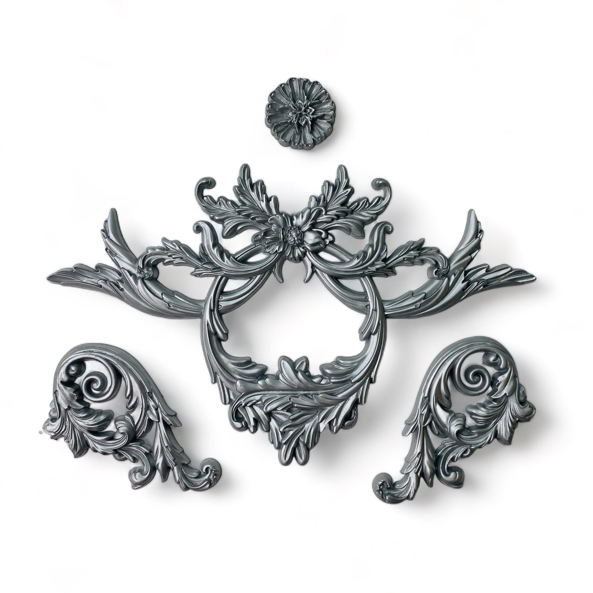Silver colored castings of ornate scrolling accent pieces and a medallion are against a white background.