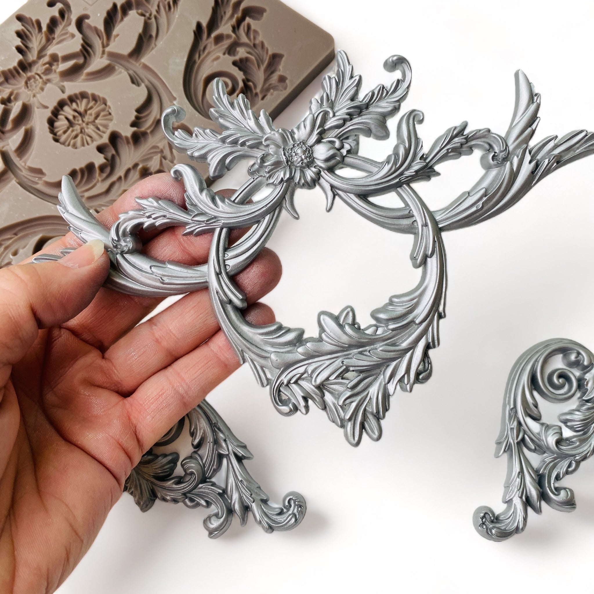 A brown silicone mold and a hand holding a medallion piece of silver colored castings of ornate scrolling accent pieces and a medallion are against a white background.