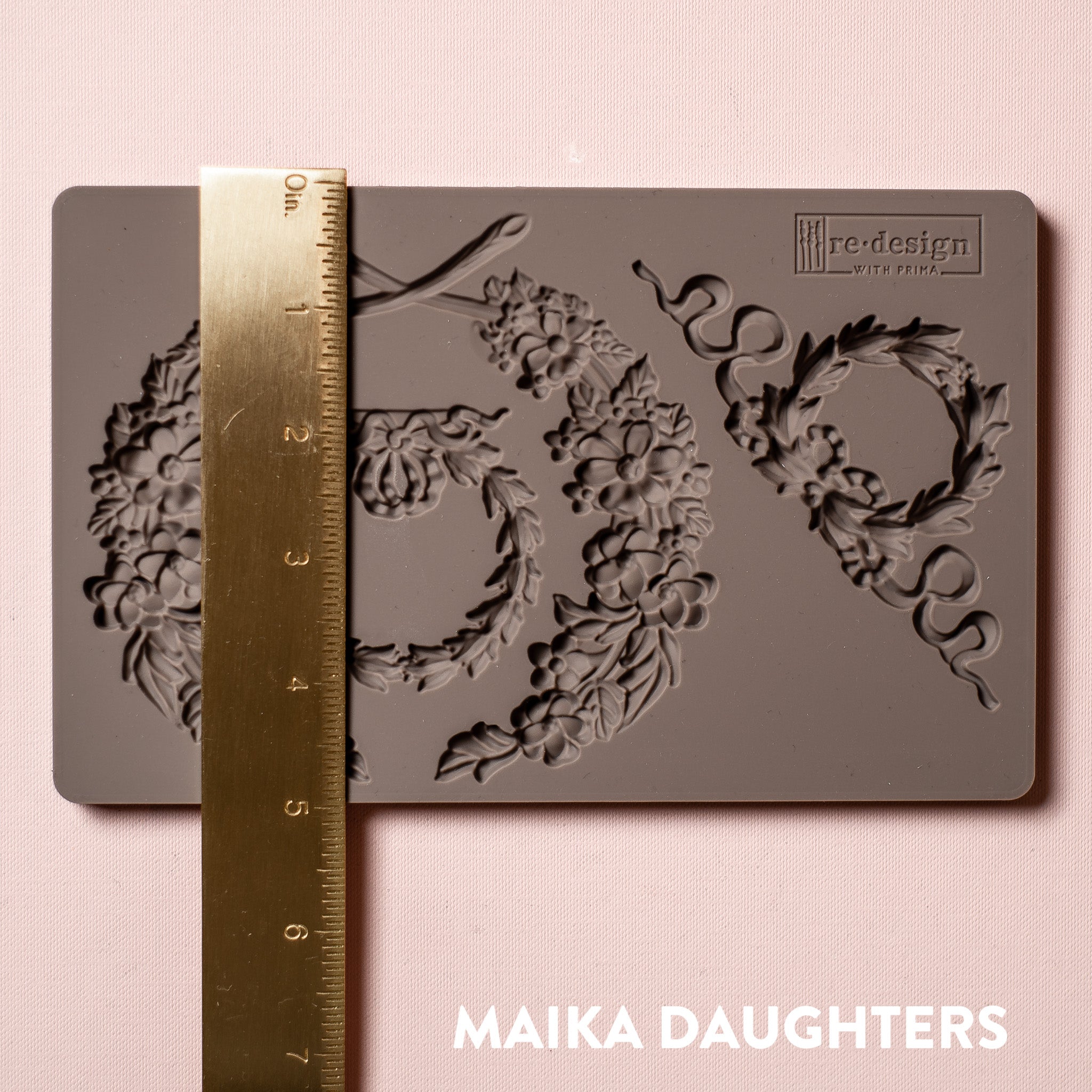 A brown silicone mold of Redesign with Prima's Laurel Wreath is against a light pink background. A gold ruler reading 5 inches sits on top of the mold.