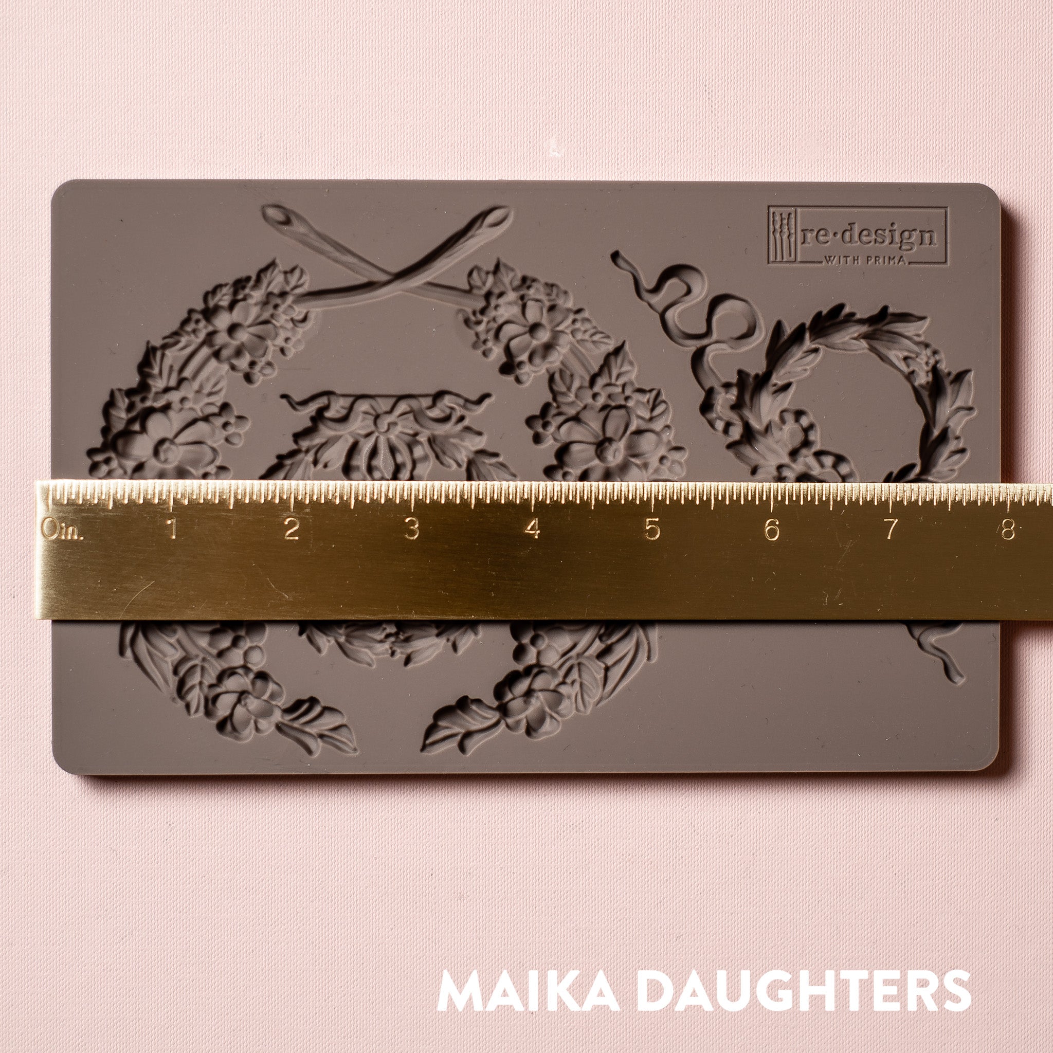A brown silicone mold of Redesign with Prima's Laurel Wreath is against a light pink background. A gold ruler reading 8 inches sits on top of the mold.