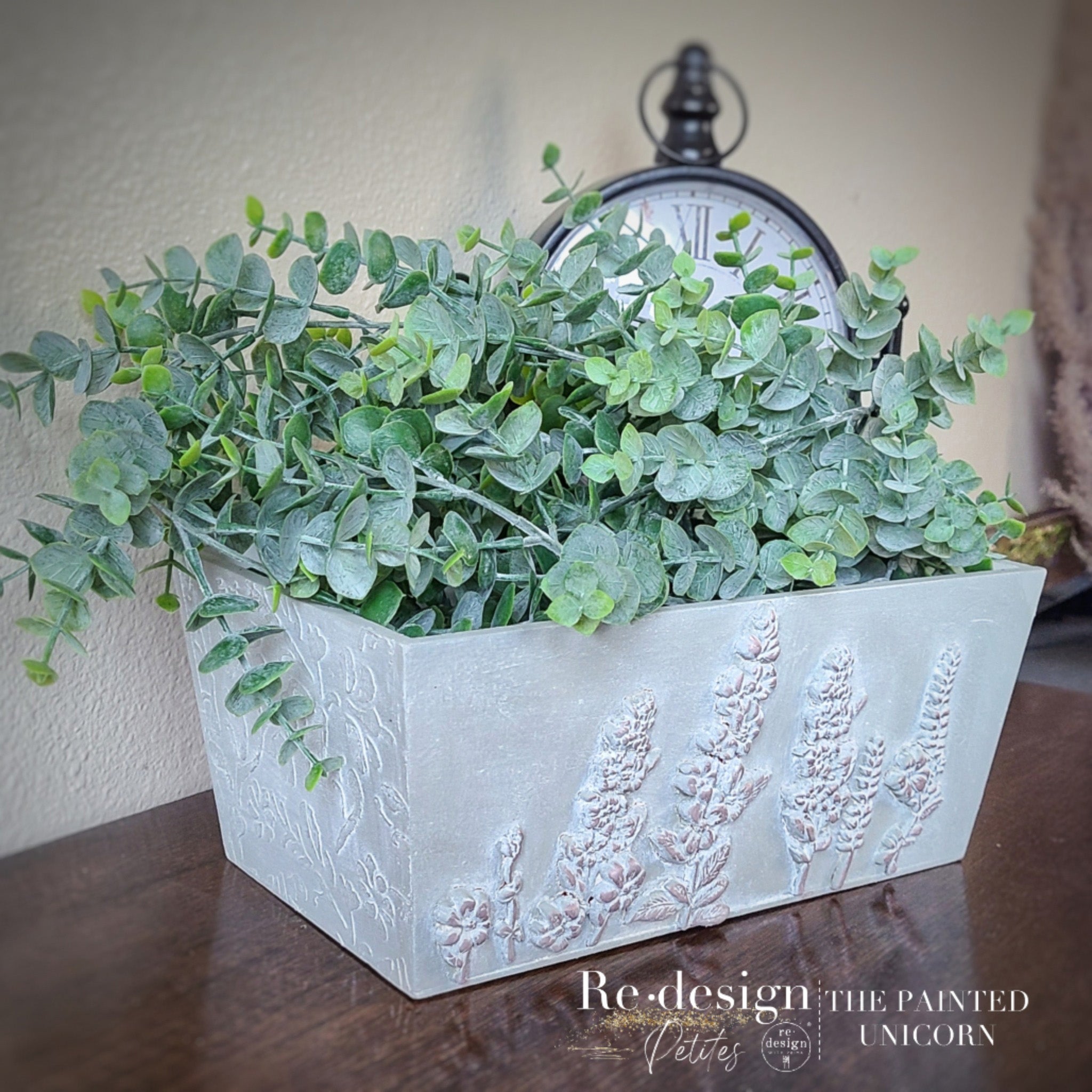 A rectangular planter refurbished by The Painted Unicorn is painted white and features ReDesign with Prima's Lavender Harvest silicone mold castings on it. Green eucalyptus is in the planter.