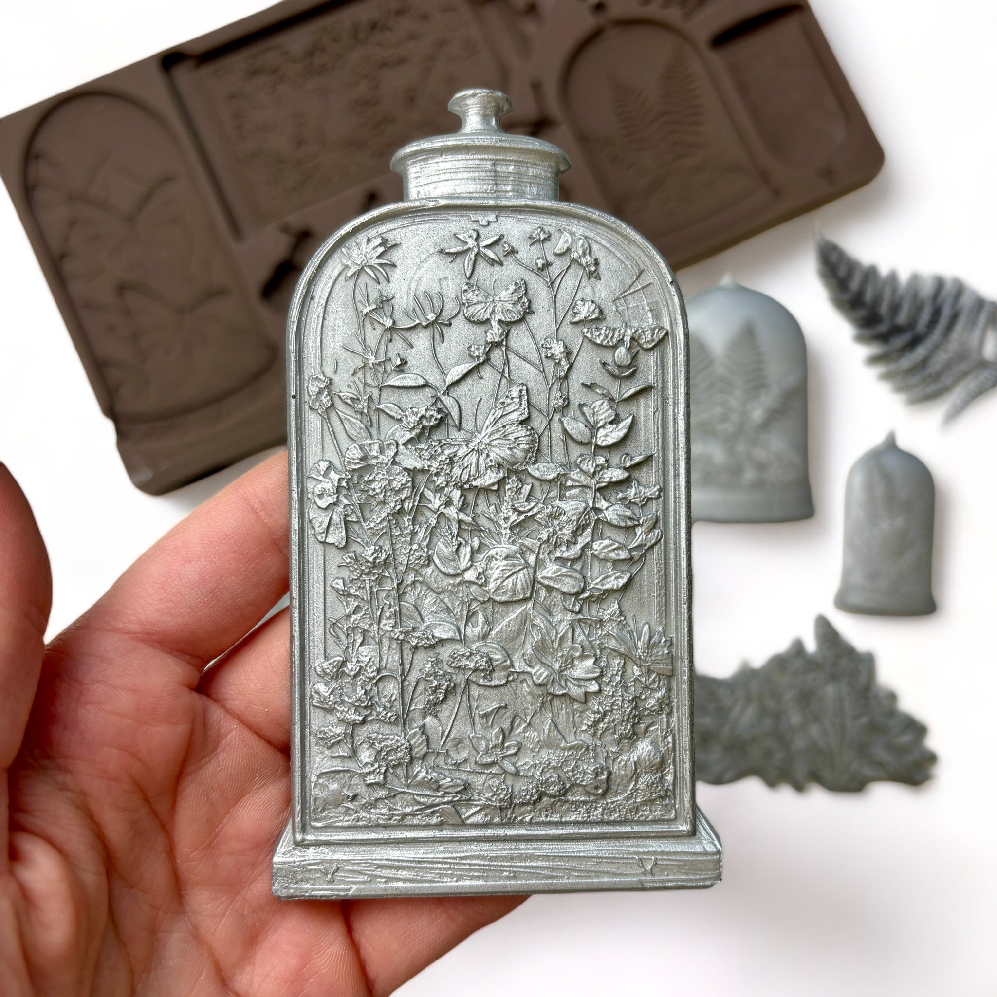 A brown silicone mold and silver colored castings of ferns, insects, mushrooms, and vintage glass cloche containers are against a white background. A hand is shown holding one of the cloches.