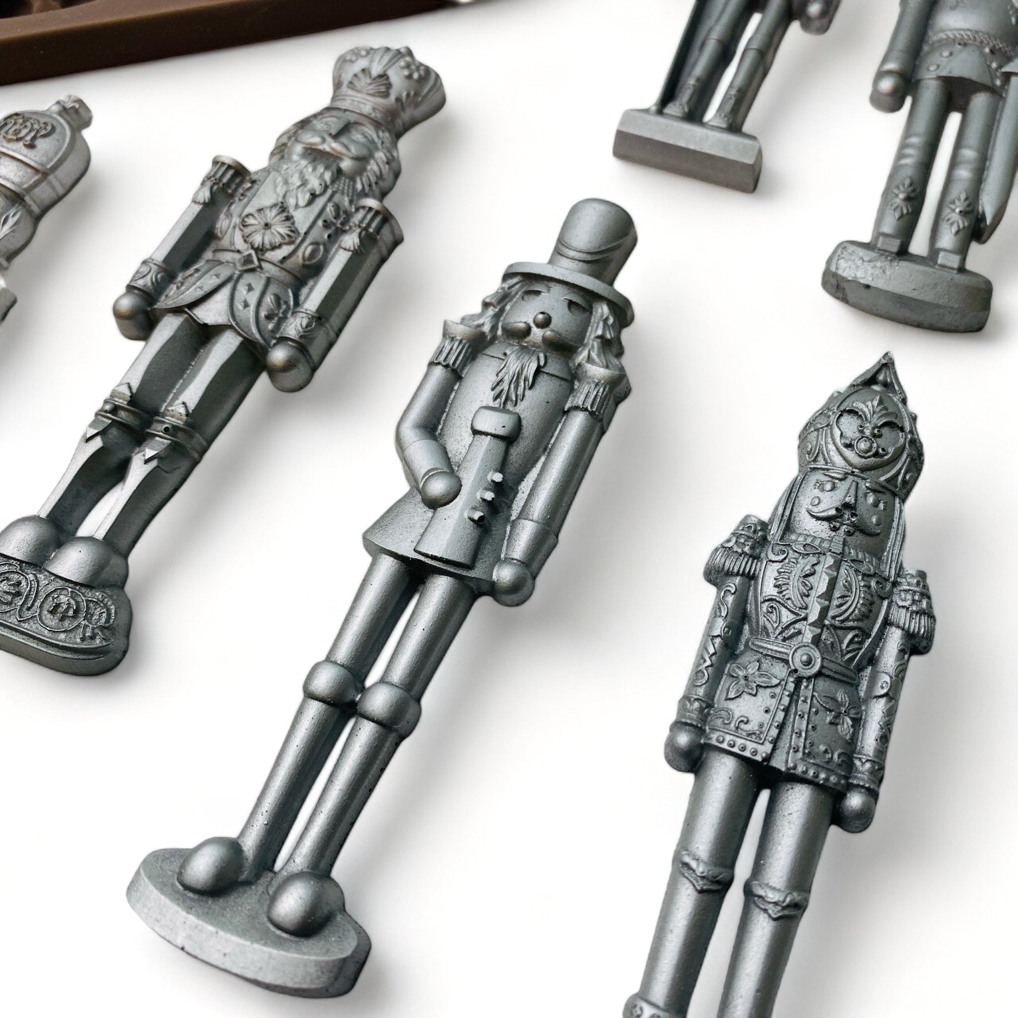 Close-up of a brown silicone mold and silver colored castings of 6 ornately decorated nutcrackers are against a white background.