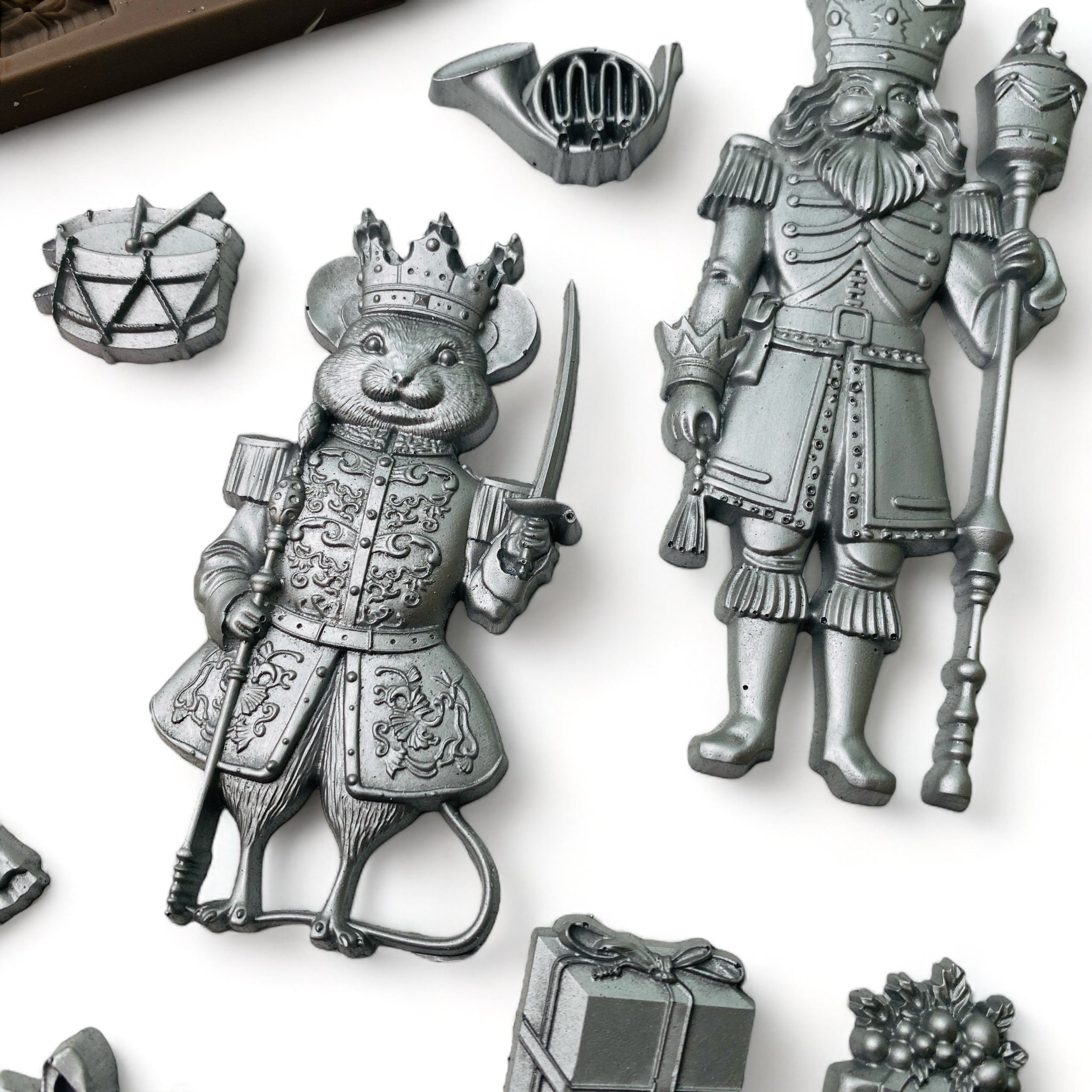 Close-up of a brown silicone mold and silver colored castings of pieces and characters from The Nutcracker are against a white background.