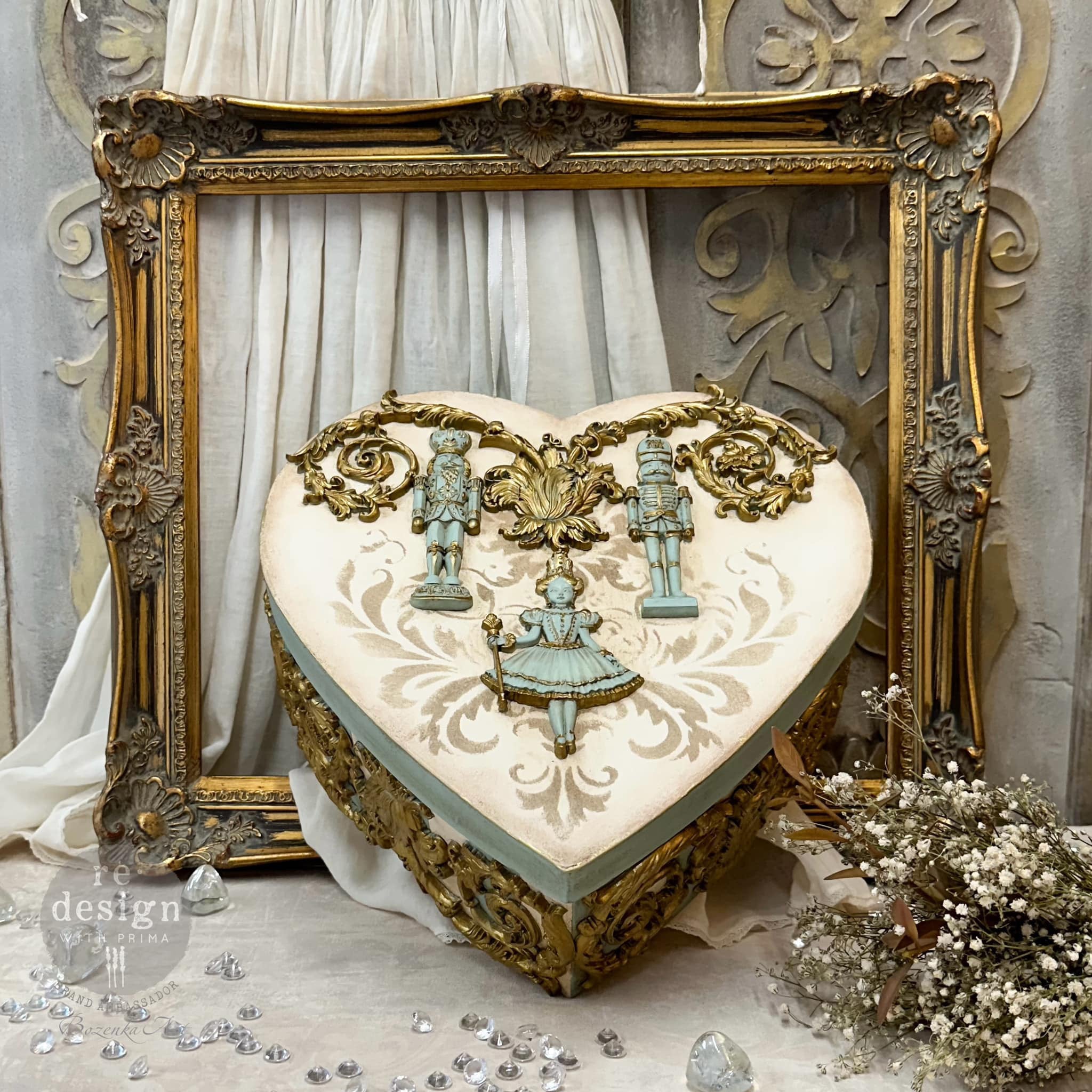 A heart-shaped box in front of a gold picture frame features gold and patina colored castings of ReDesign with Prima's Nutcracker Party silicone mold.