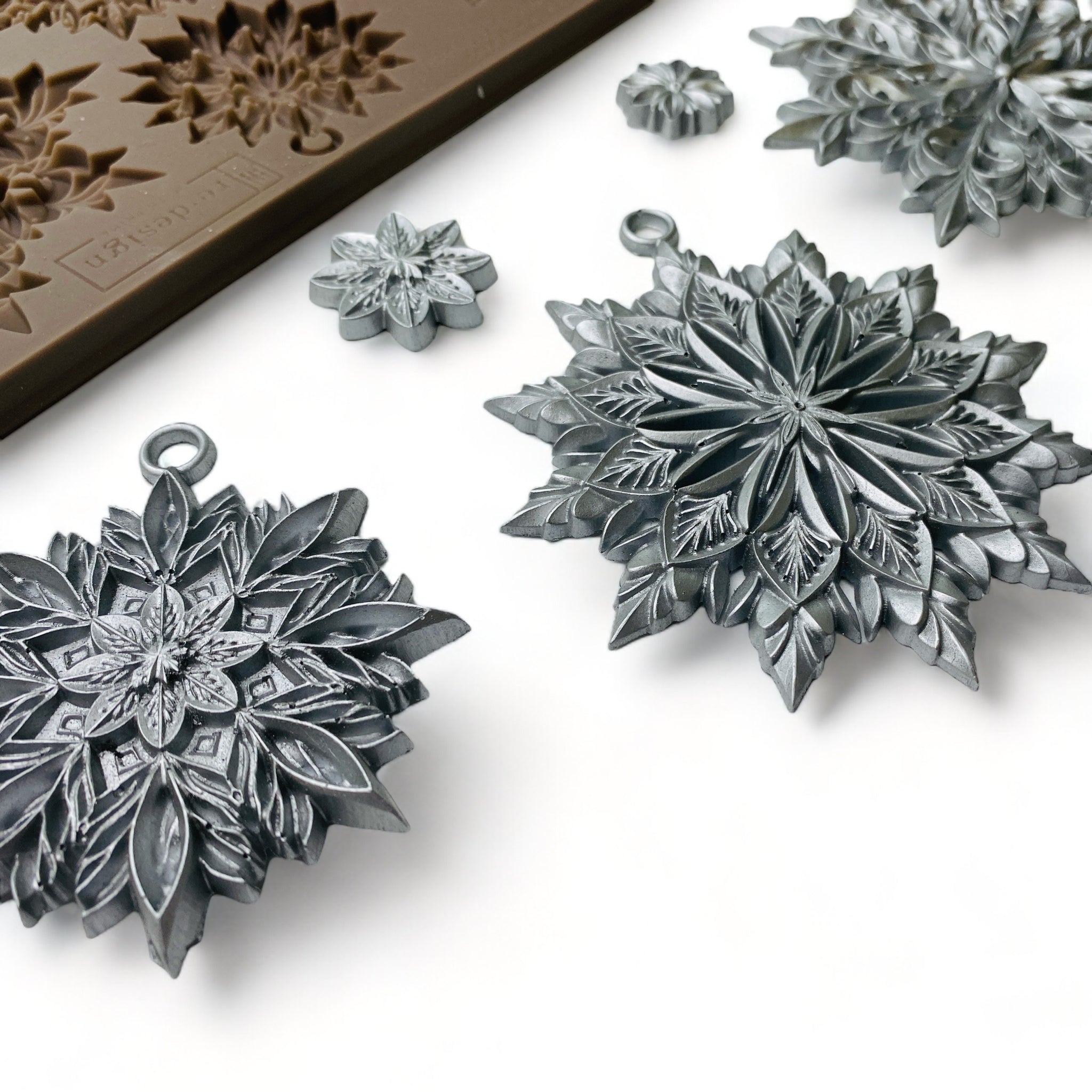 Close-up of a brown silicone mold and silver colored castings of 3 large, and 5 smaller ornate snowflakes are against a white background.