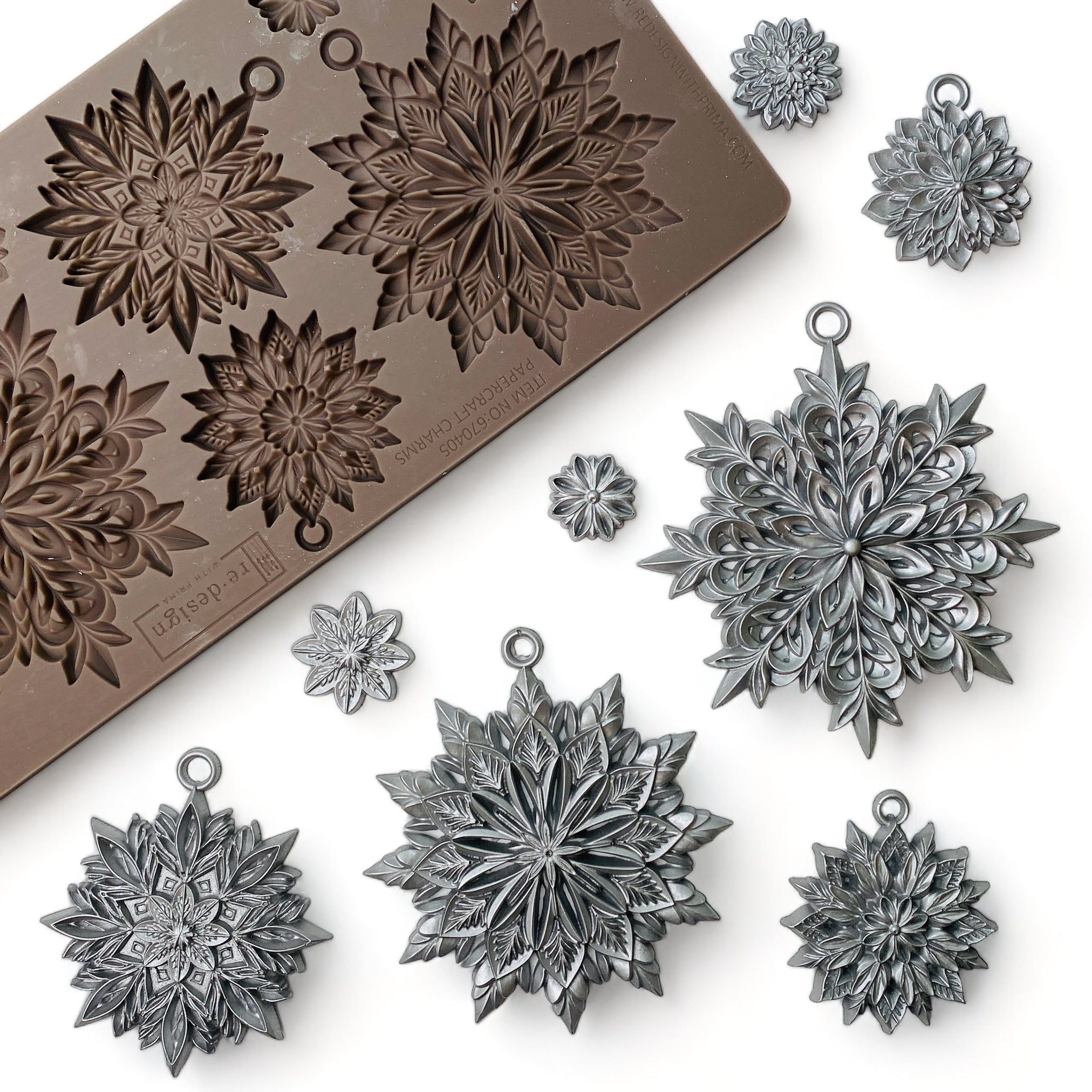 A brown silicone mold and silver colored castings of 3 large, and 5 smaller ornate snowflakes are against a white background.