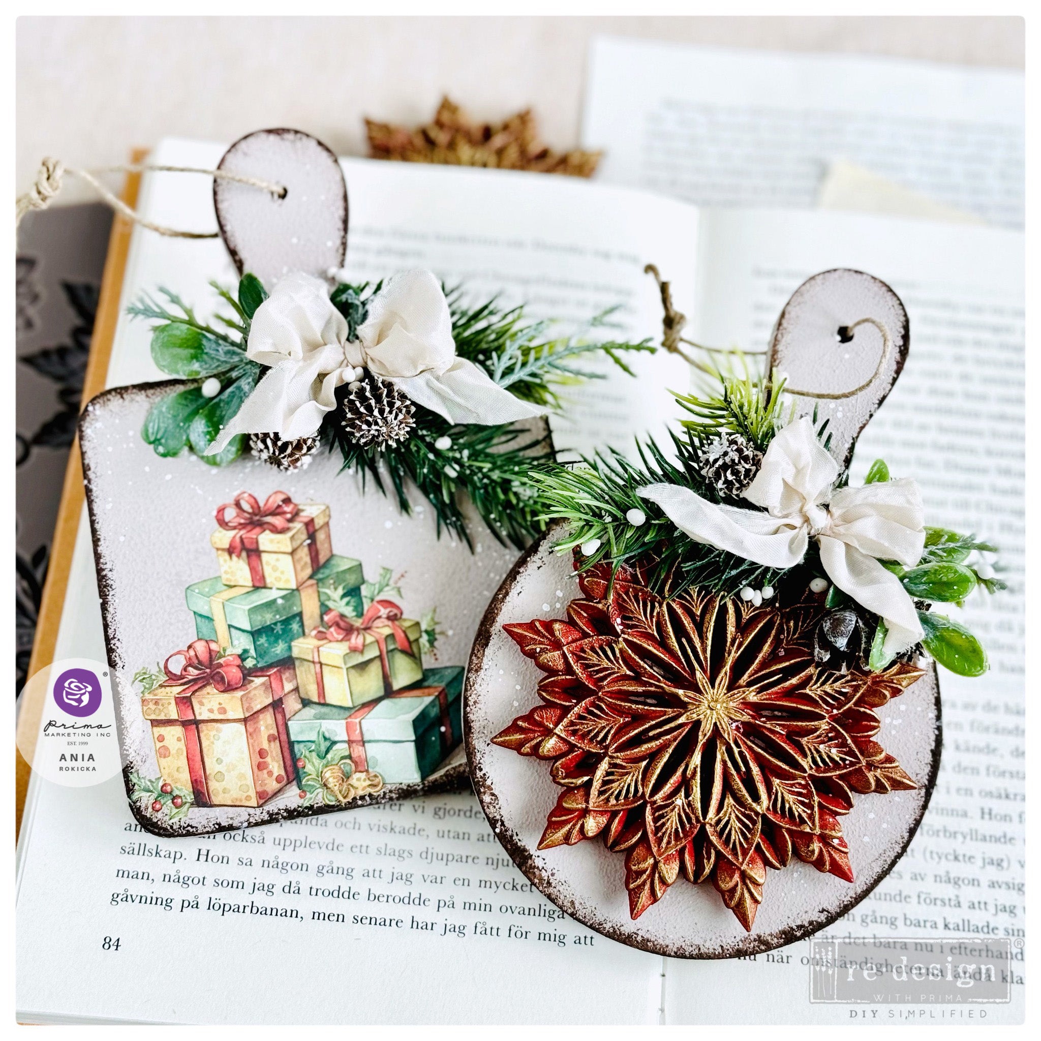 Two white glitter metal ornaments are against an open book. One of the ornaments features a red and gold painted casting from ReDesign with Prima's Papercraft Charms silicone mold.