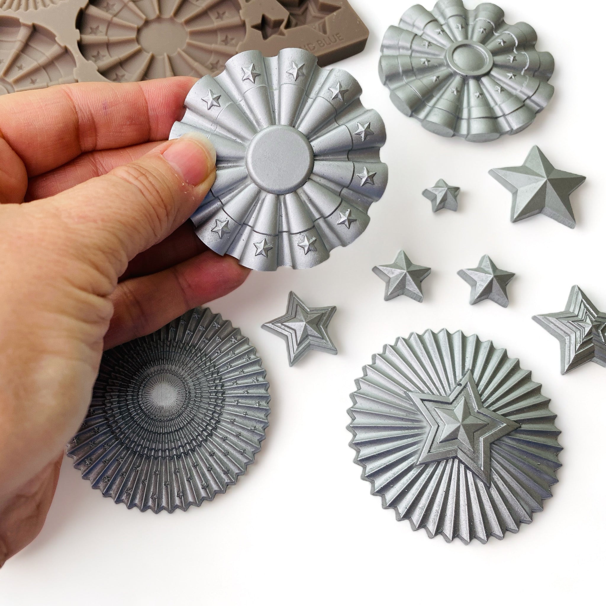 A brown silicone mold and silver colored castings of ReDesign with Prima's Red, White, and Blue silicone mold are against a white background. A hand is shown holding one of the round ribbon designs from the mold.