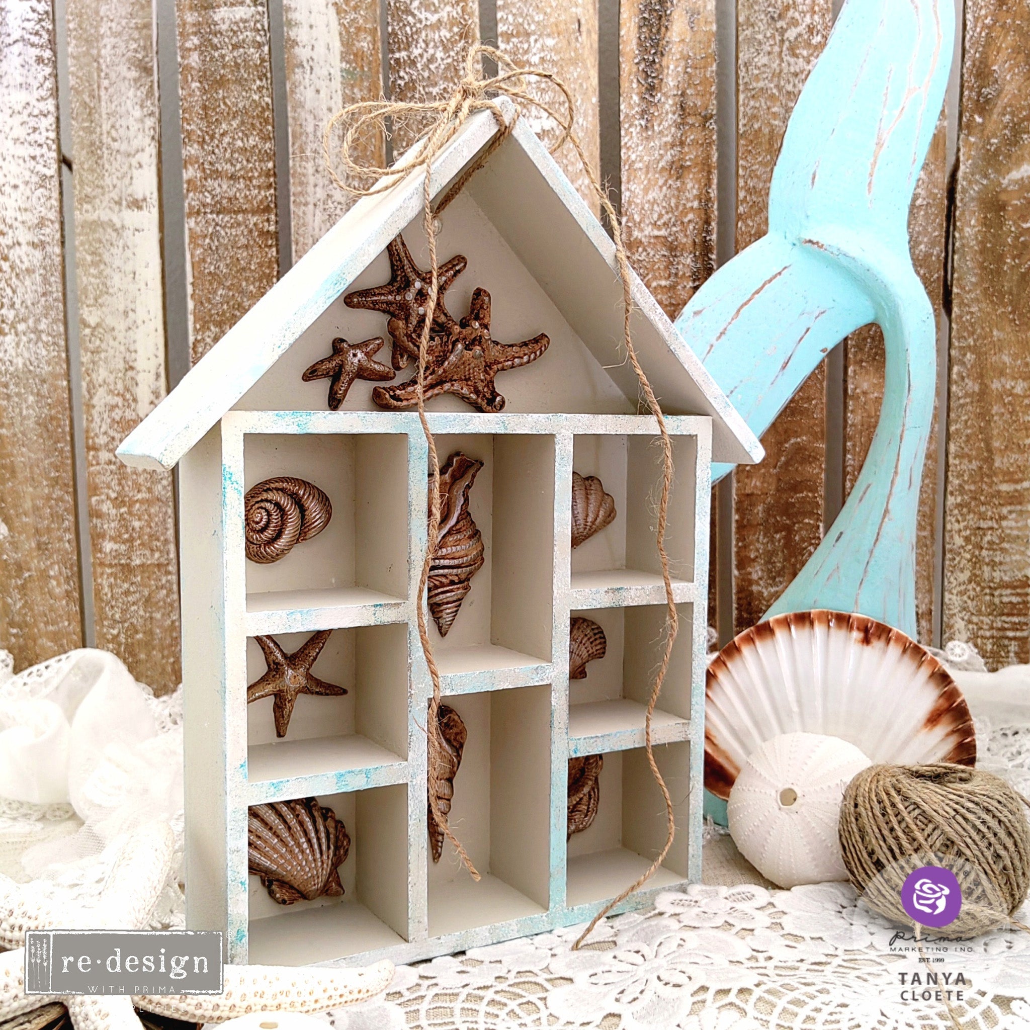 A small house-shaped hanging shelving box created by Tanya Cloete is painted white and features ReDesign with Prima's Reef Elegance silicone mold castings in bronze color inside each cubby hole.