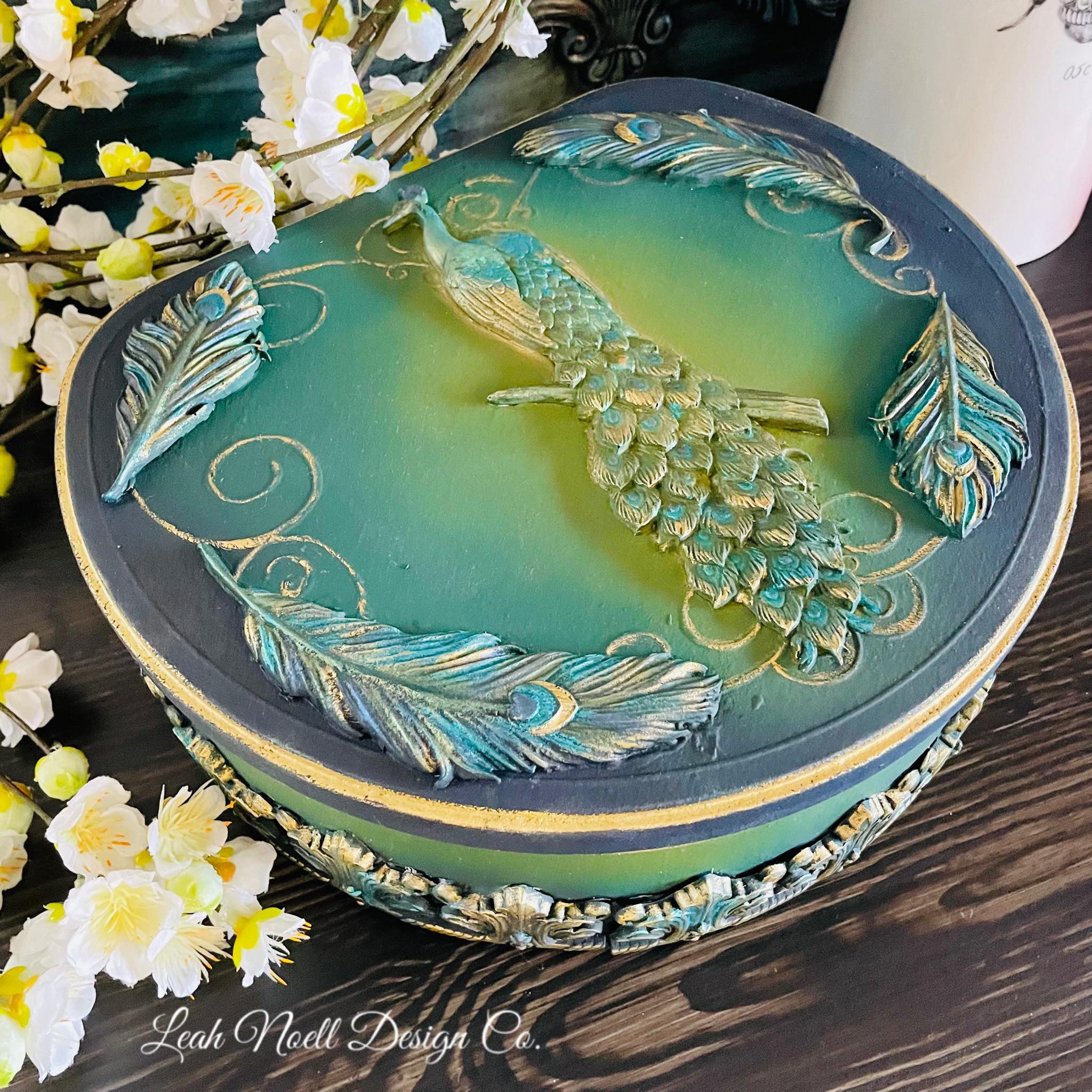 A hat box created by Leah Noell Design Company is painted a blend of blue, green, and yellow and features castings of ReDesign with Prima's Regal Peacock silicone mold on it.