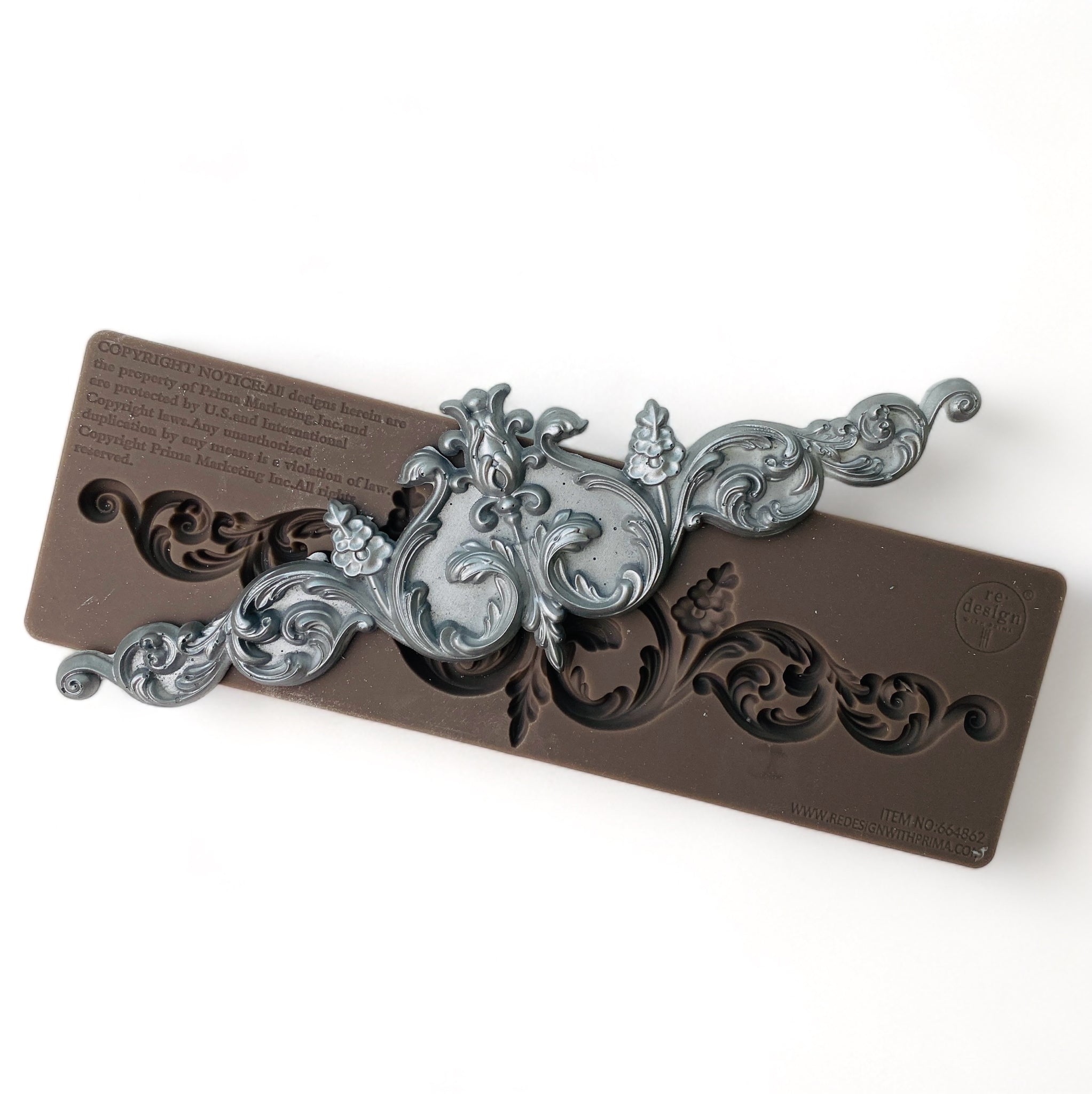 A brown silicone mold and silver colored casting of an ornate scroll design are against a white background.