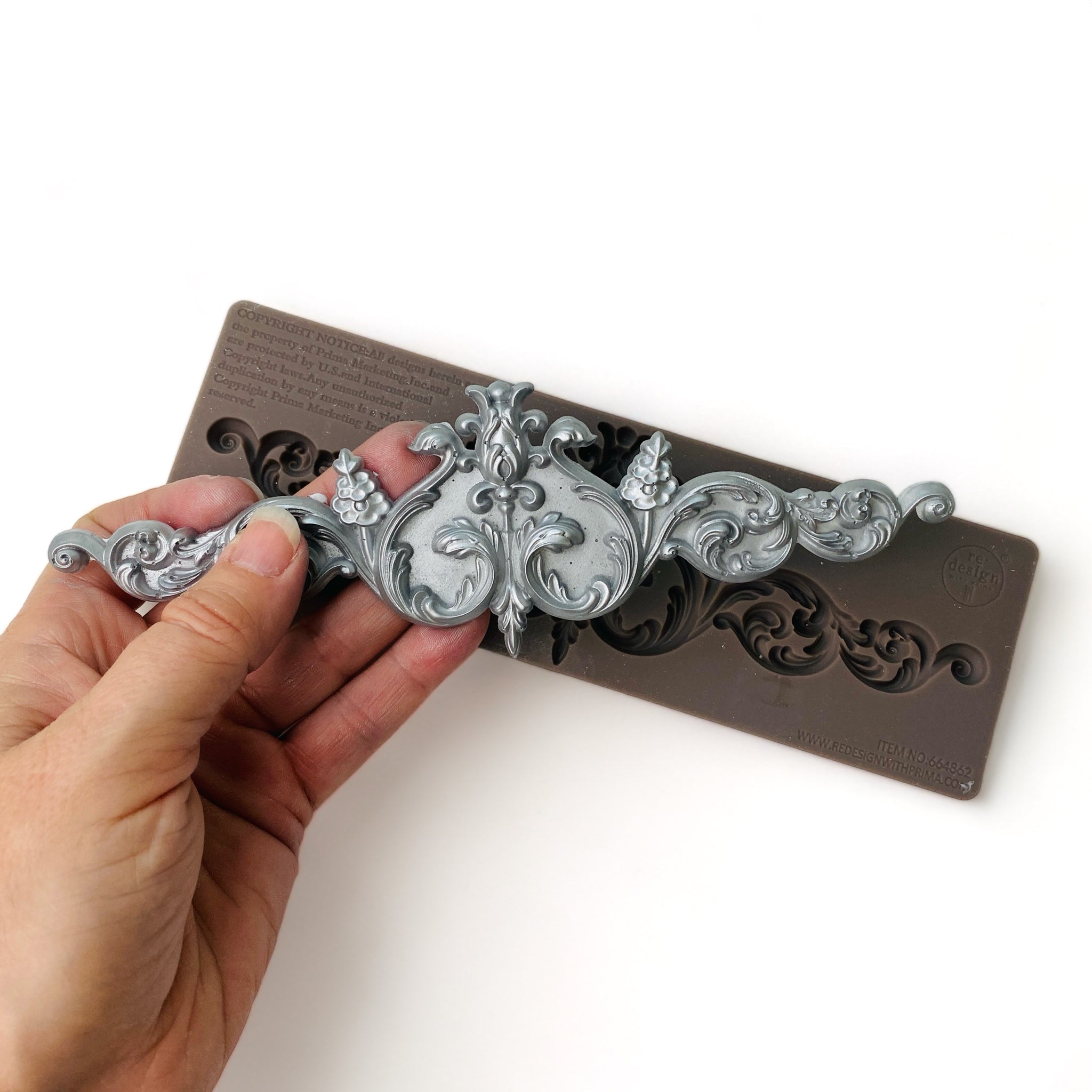 A brown silicone mold and a hand holding a silver colored casting of an ornate scroll design are against a white background.