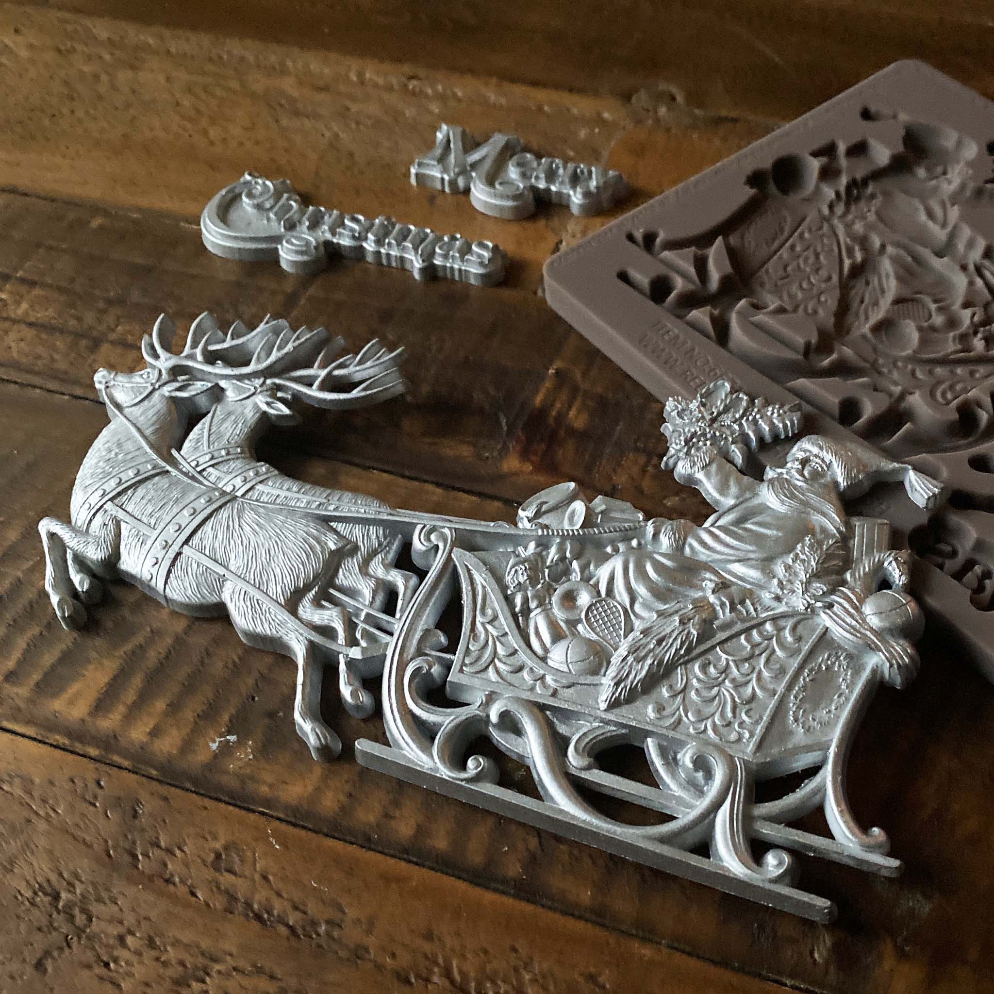 Silver colored castings of ReDesign with Prima's Santa's Sleigh sit on their brown silicone mould against a dark wood background.