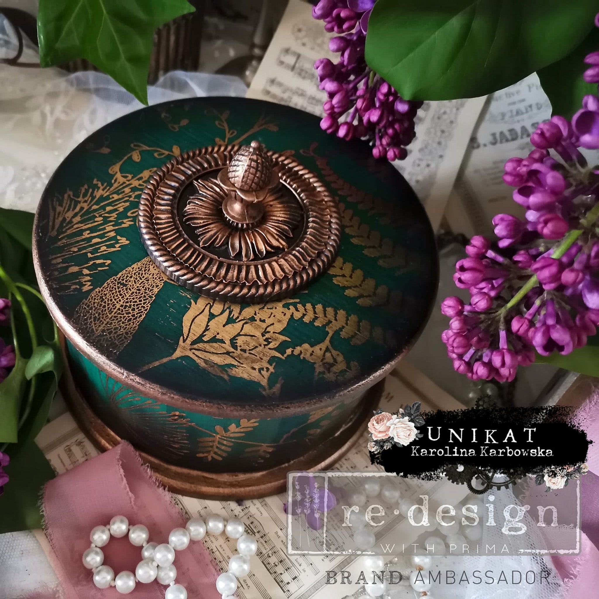 A cylindrical trinket box created by Unikat Karolina Karbowska is painted dark green with gold plants stenciling and features ReDesign with Prima's Soho Haven silicone mold on the top with a knob.