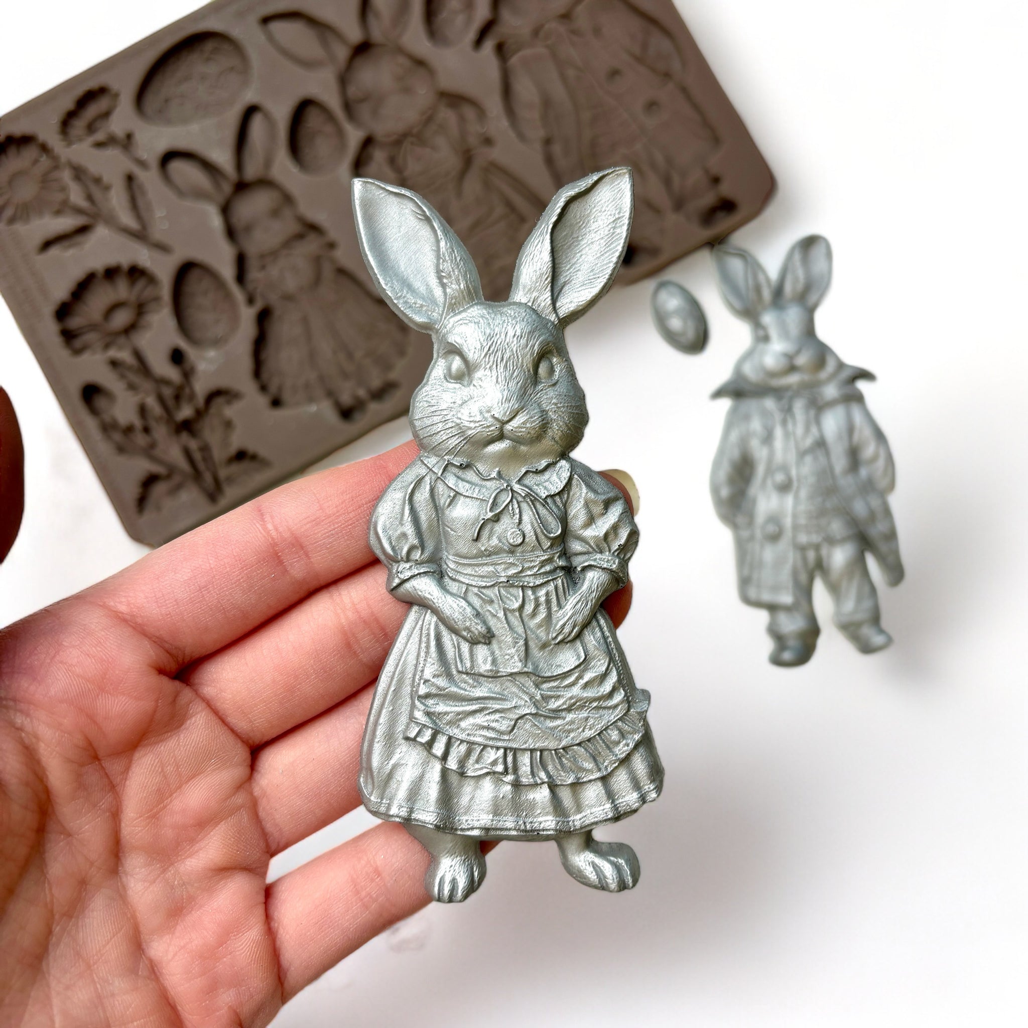 A brown silicone mold and silver colored castings of 3 cute bunnies in clothes, 3 flowers, and 4 Easter eggs are against a white background. A hand is shown holding one of the bunny castings.