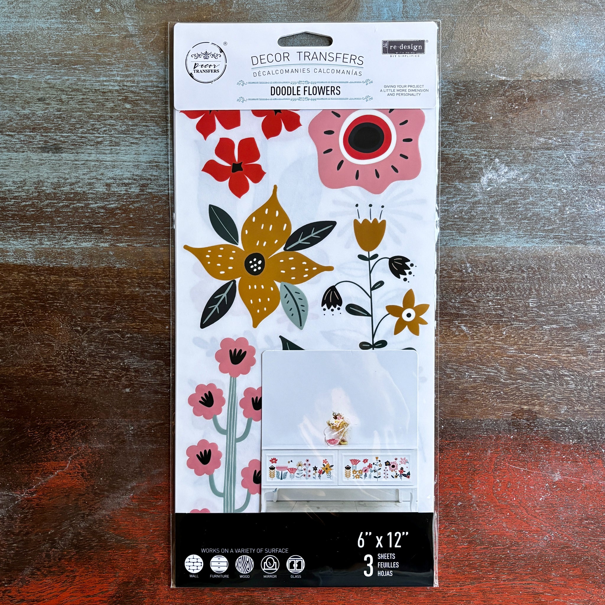 A package of ReDesign with Prima's Doodle Flowers small transfer is against a dark wood background.