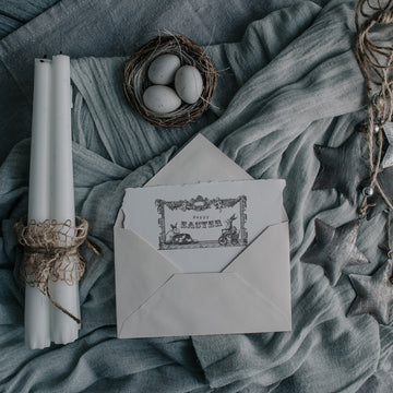 Three white candlesticks, a small nest with 3 eggs, and a handmade card peeking out of an envelope that features 1 of ReDesign with Prima's Easter Blessings stamp are against some bundled up light blue material. 