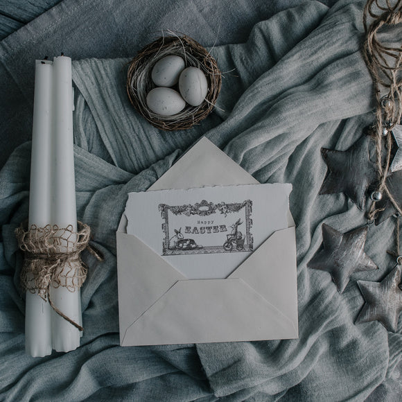 Three white candlesticks, a small nest with 3 eggs, and a handmade card peeking out of an envelope that features 1 of ReDesign with Prima's Easter Blessings stamp are against some bundled up light blue material. 