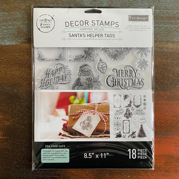A package of ReDesign with Prima's Santa's Helper Decor Stamps is against a dark wood background.
