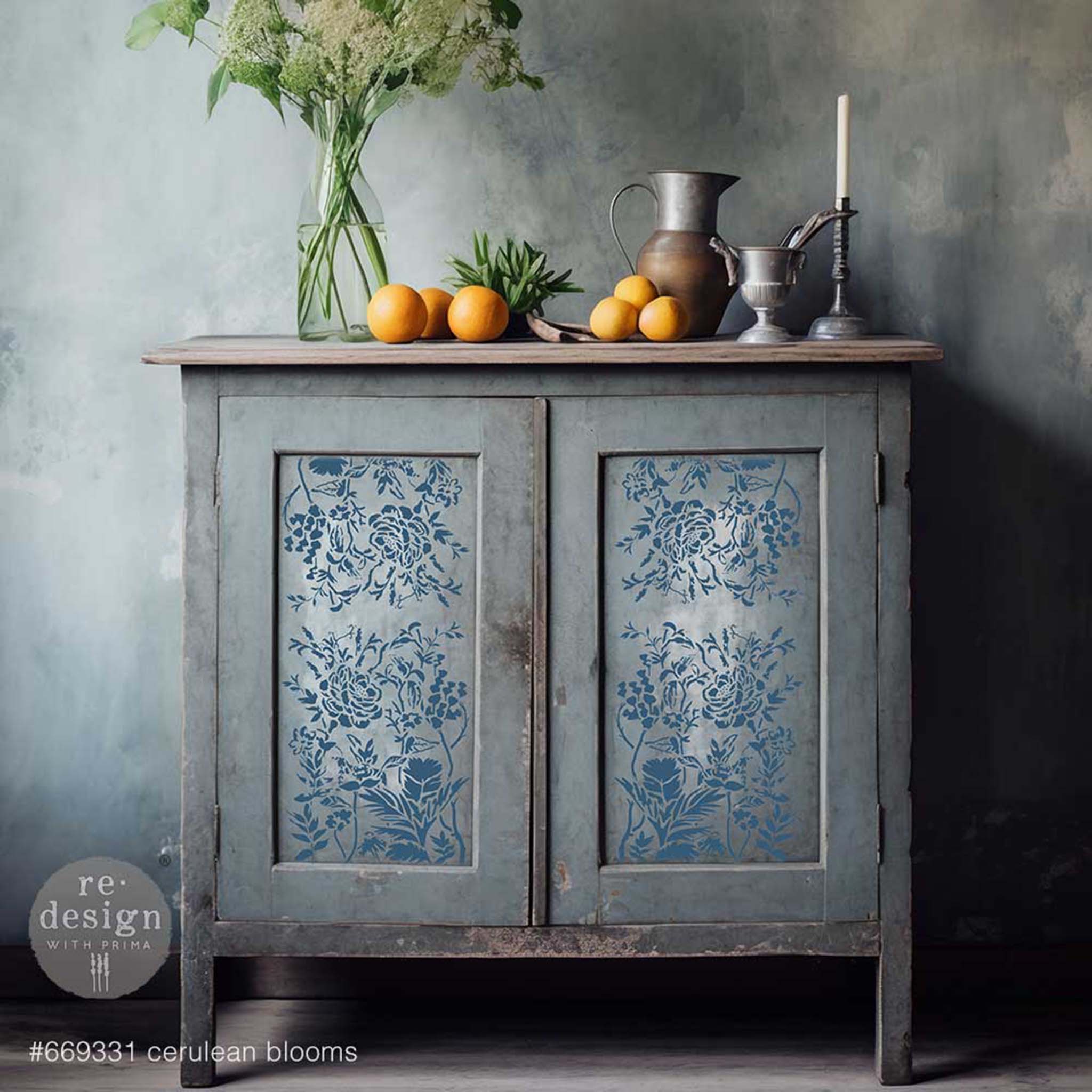 A vintage small buffet console is painted grey and feautres ReDesign with Prima's Cerulean Blooms stencil in blue inside its 2 door inlays.