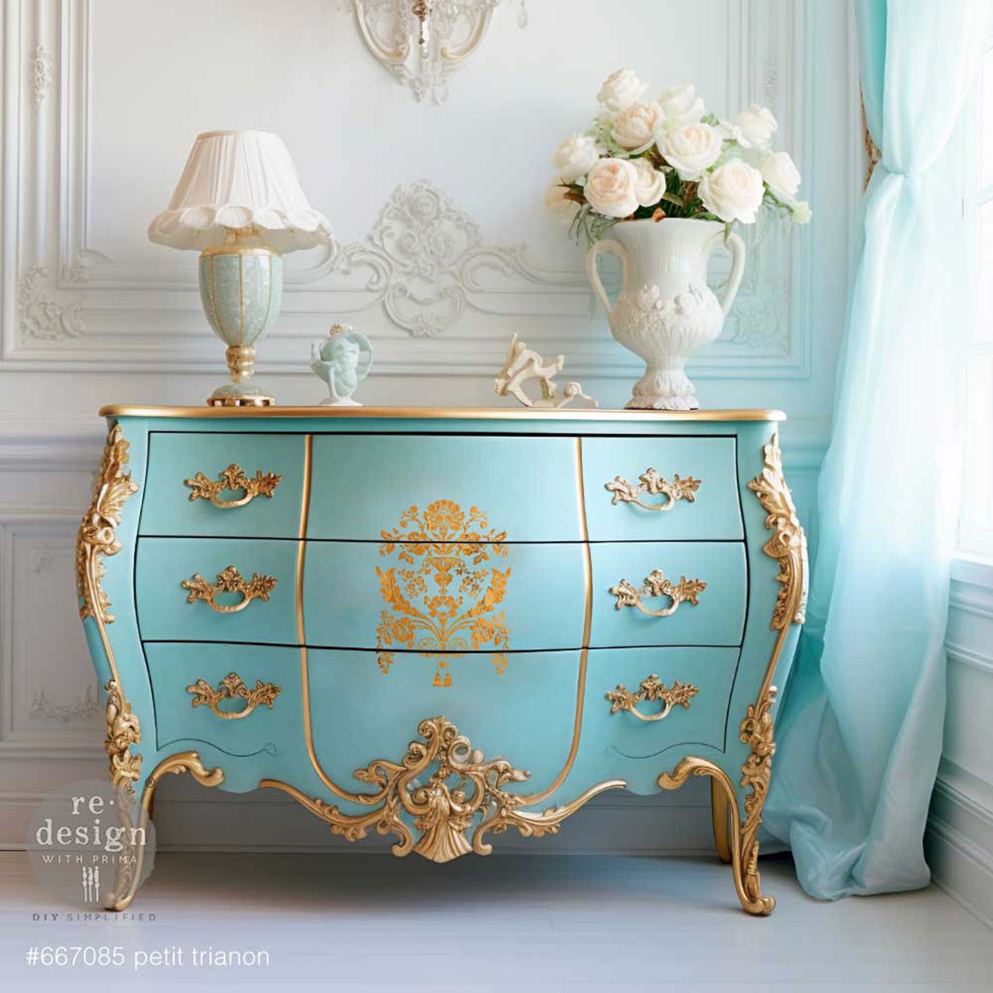 A vintage Bombay dresser is painted light teal with gold accents and features ReDesign with Prima's Petit Trianon stencil in gold in the center of the drawers.