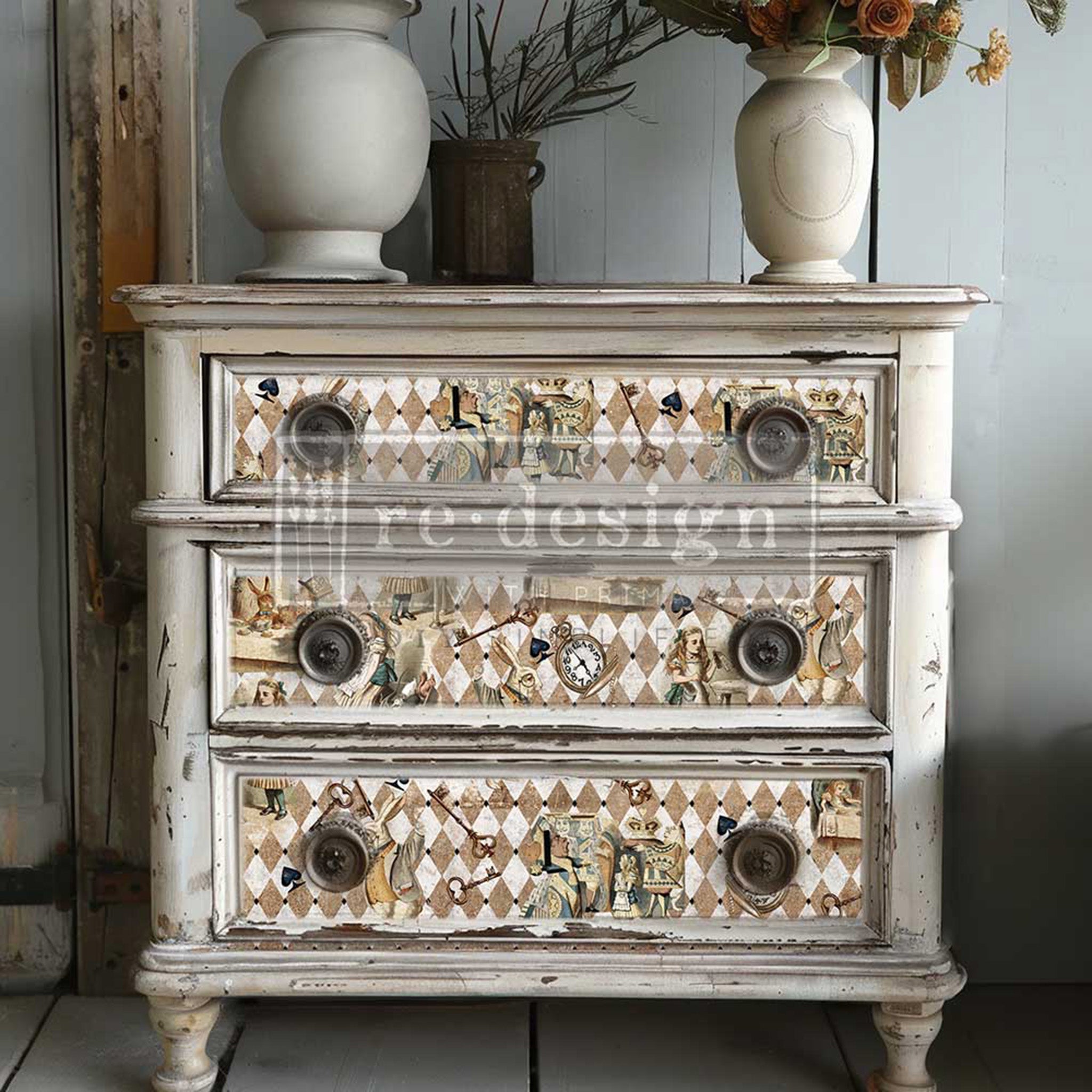 A vintage 3-drawer dresser is painted distressed white and features ReDesign with Prima's Mad Hatter's Muse tissue paper on the drawers.