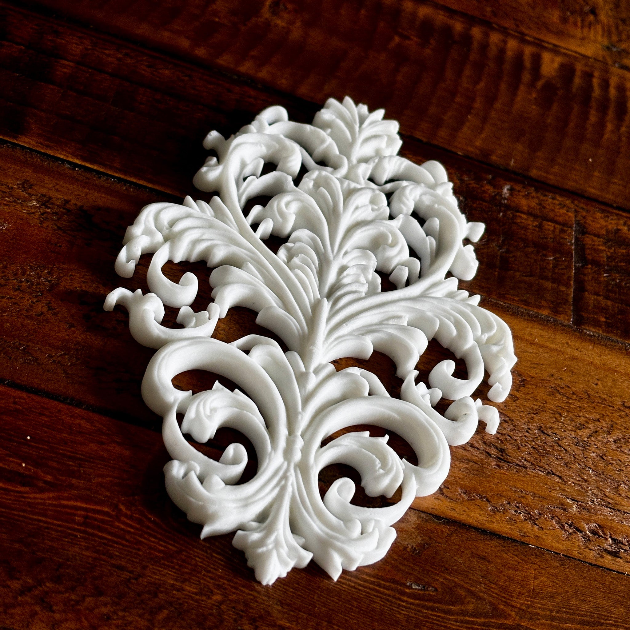 A white resin casting created from LaBlanche's Baroque Emblem silicone mold is against a dark wood background.