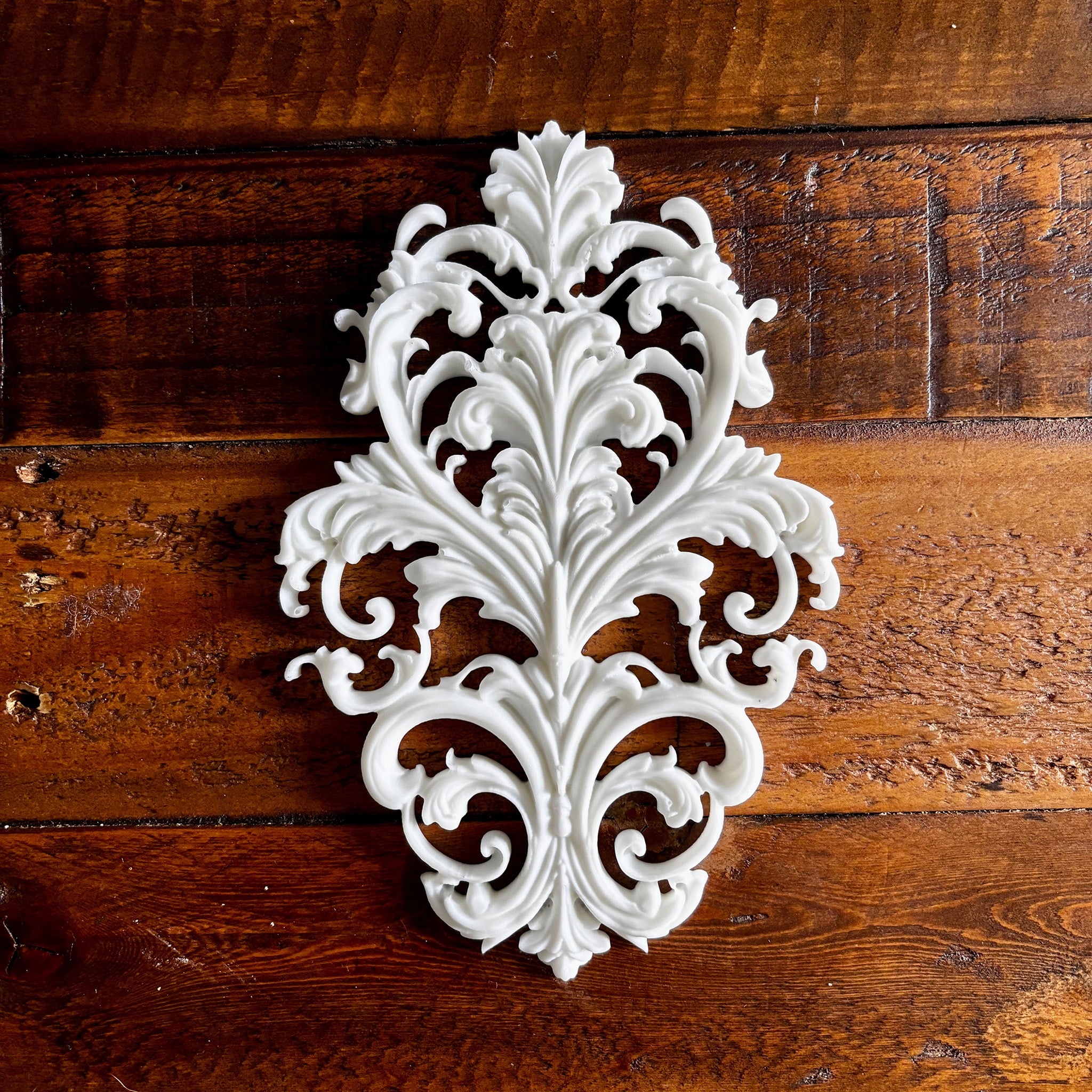 A white resin casting created from LaBlanche's Baroque Emblem silicone mold is against a dark wood background.