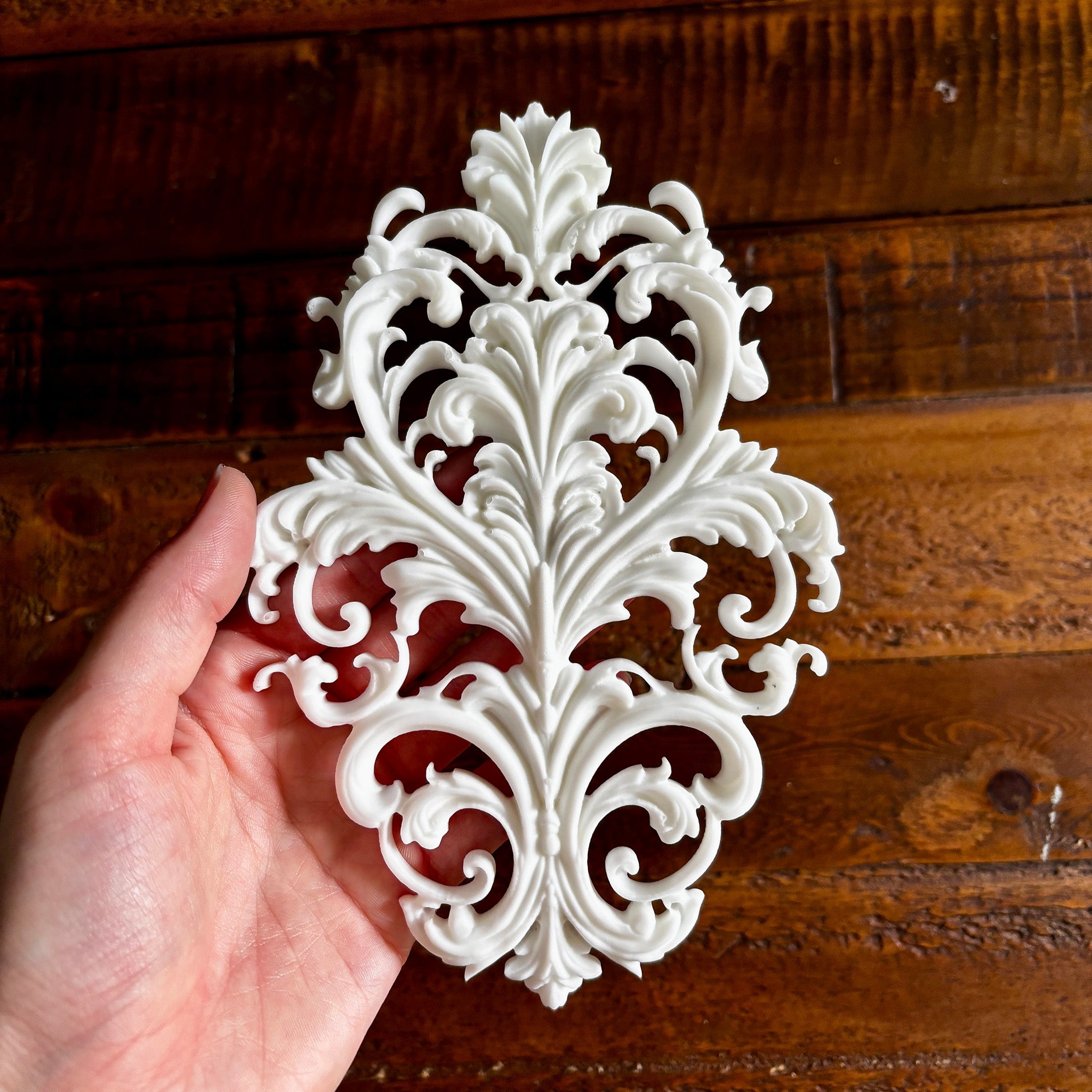 A hand is shown holding a white resin casting created from LaBlanche's Baroque Emblem silicone mold against a dark wood background. 