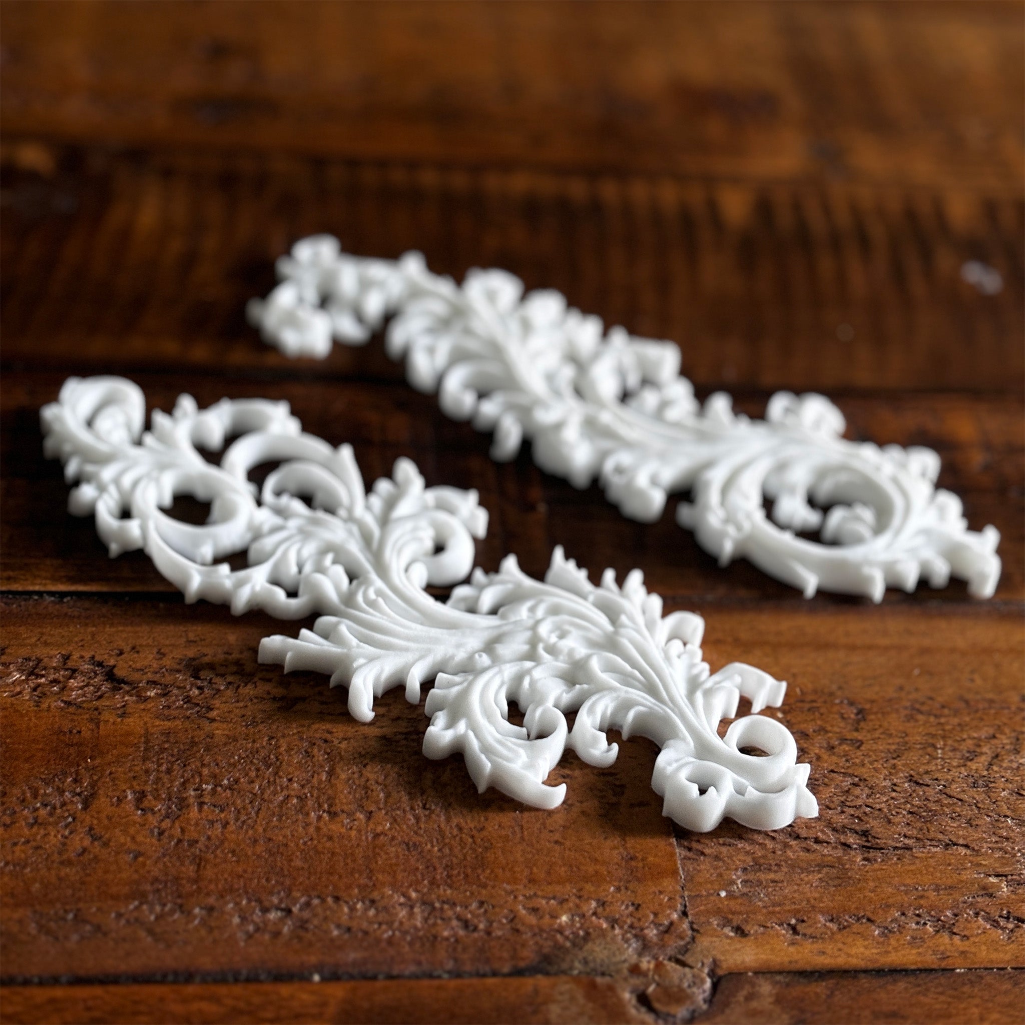 Two white resin castings of Baroque style flourishes created from LaBlanche's Curved Ornaments silicone mold are against a dark wood background.