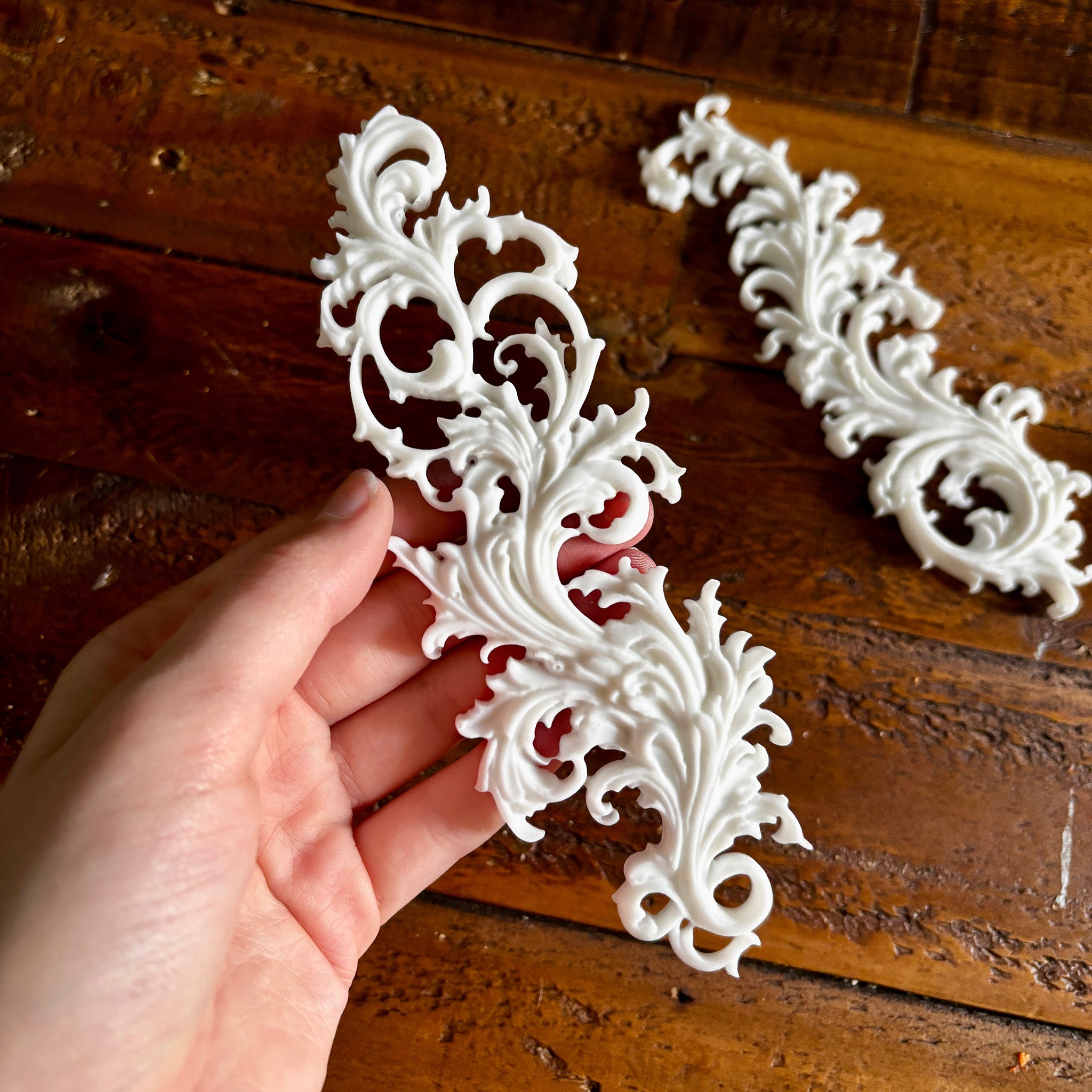 Two white resin castings of Baroque style flourishes created from LaBlanche's Curved Ornaments silicone mold are against a dark wood background. A hand is shown holding one of the castings.