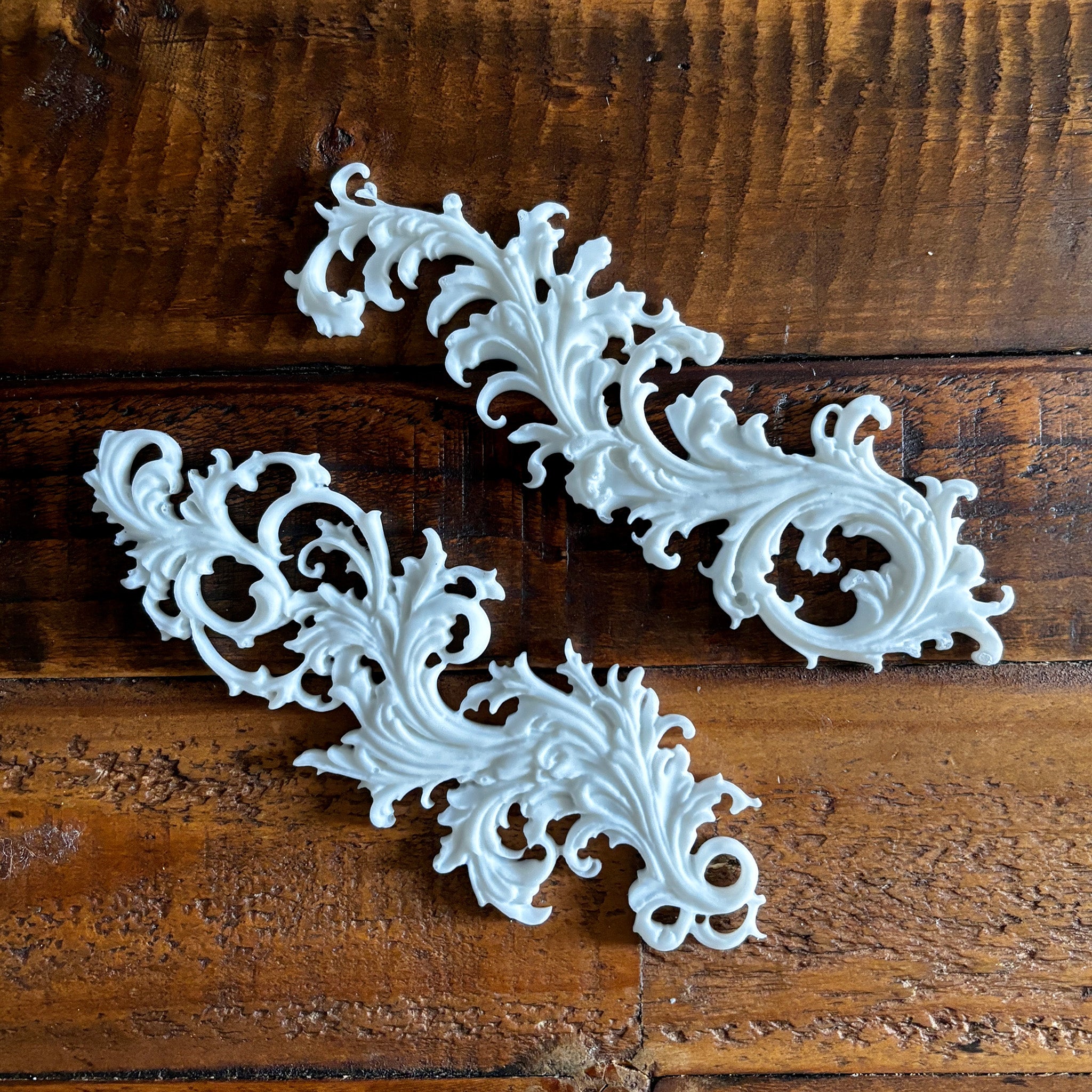 Two white resin castings of Baroque style flourishes created from LaBlanche's Curved Ornaments silicone mold are against a dark wood background.