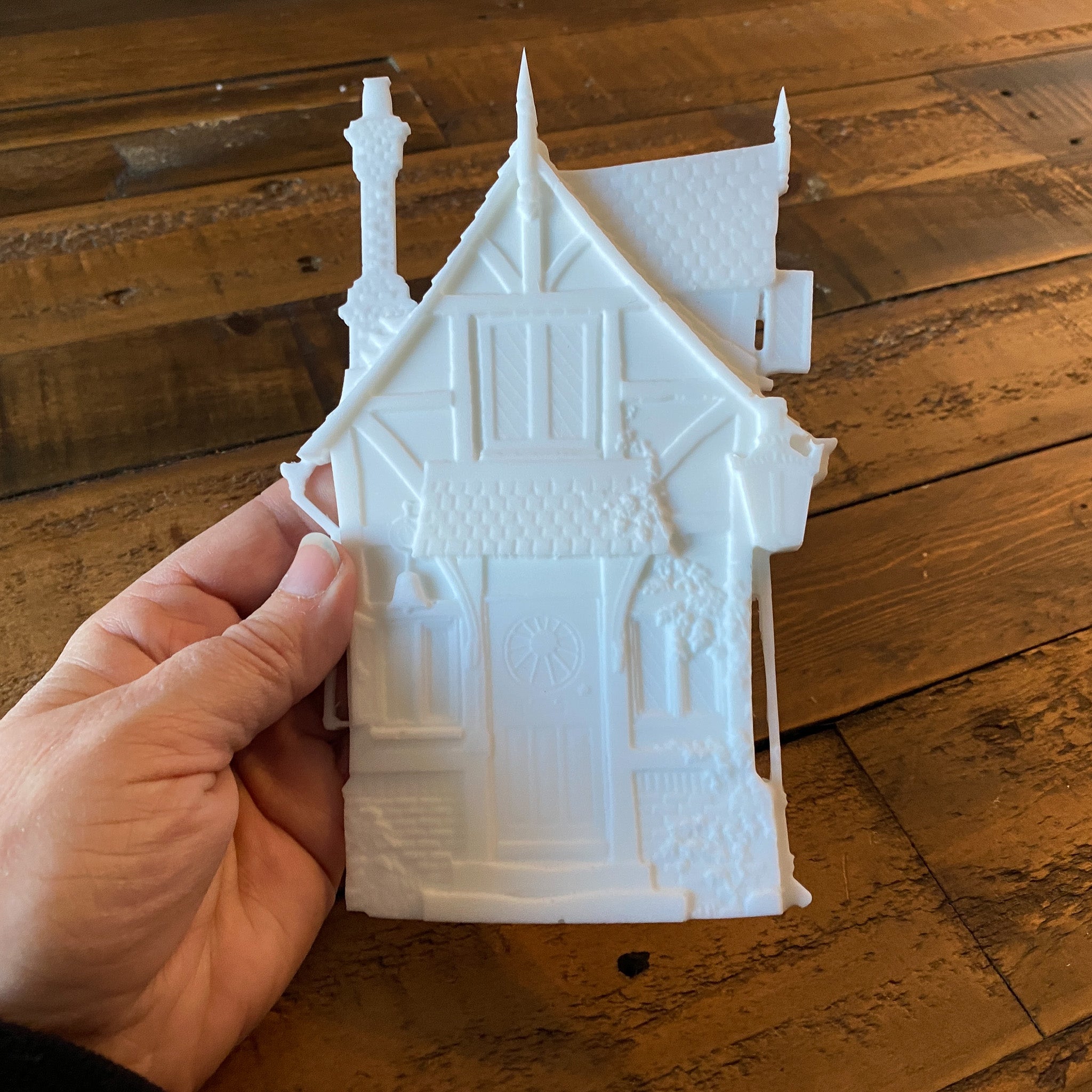 A hand holding a white resin silicone mold casting of a small Victorian style fairy house is against a wood background.