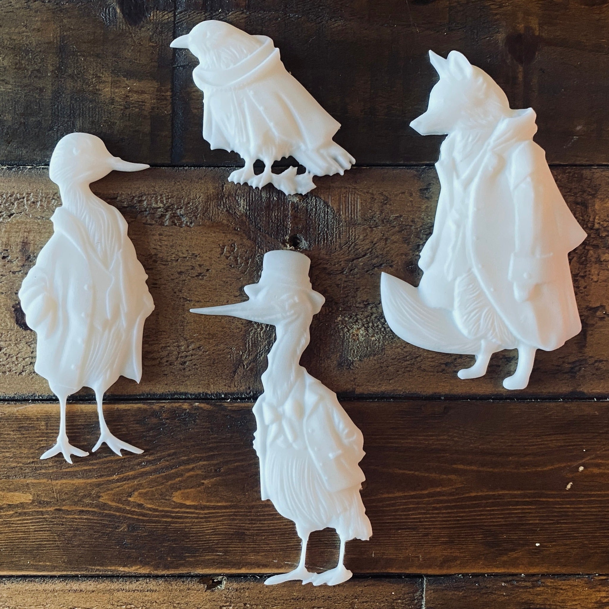 White resin castings of a fox and 3 birds all wearing vintage overcoats are against a wood background.