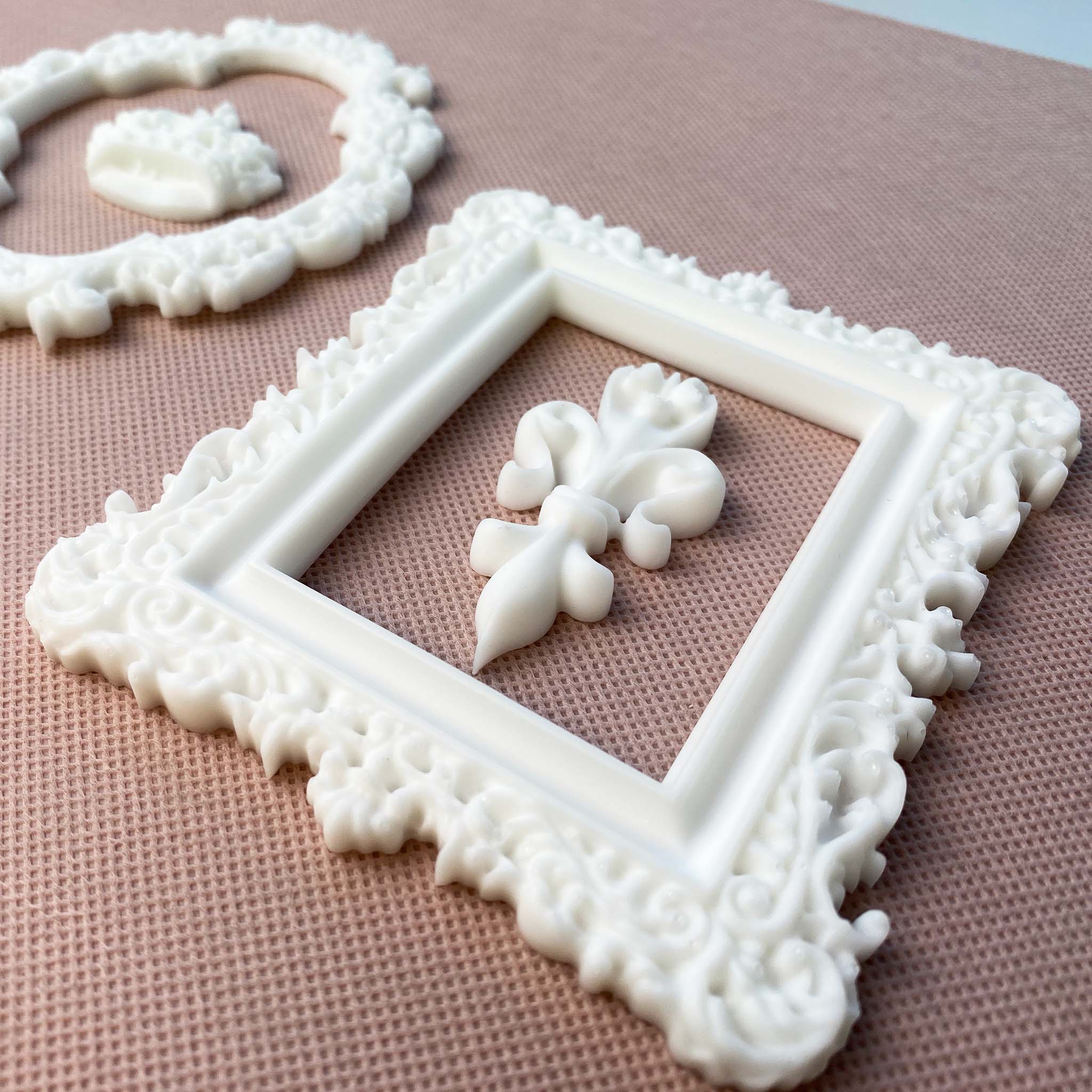 An angled view to show the thickness of white resin castings of an ornate oval and rectangle frame are against a light beige material background. Inside the oval frame is a casting of a royal crown and inside the rectangle frame is a fleur de lis.