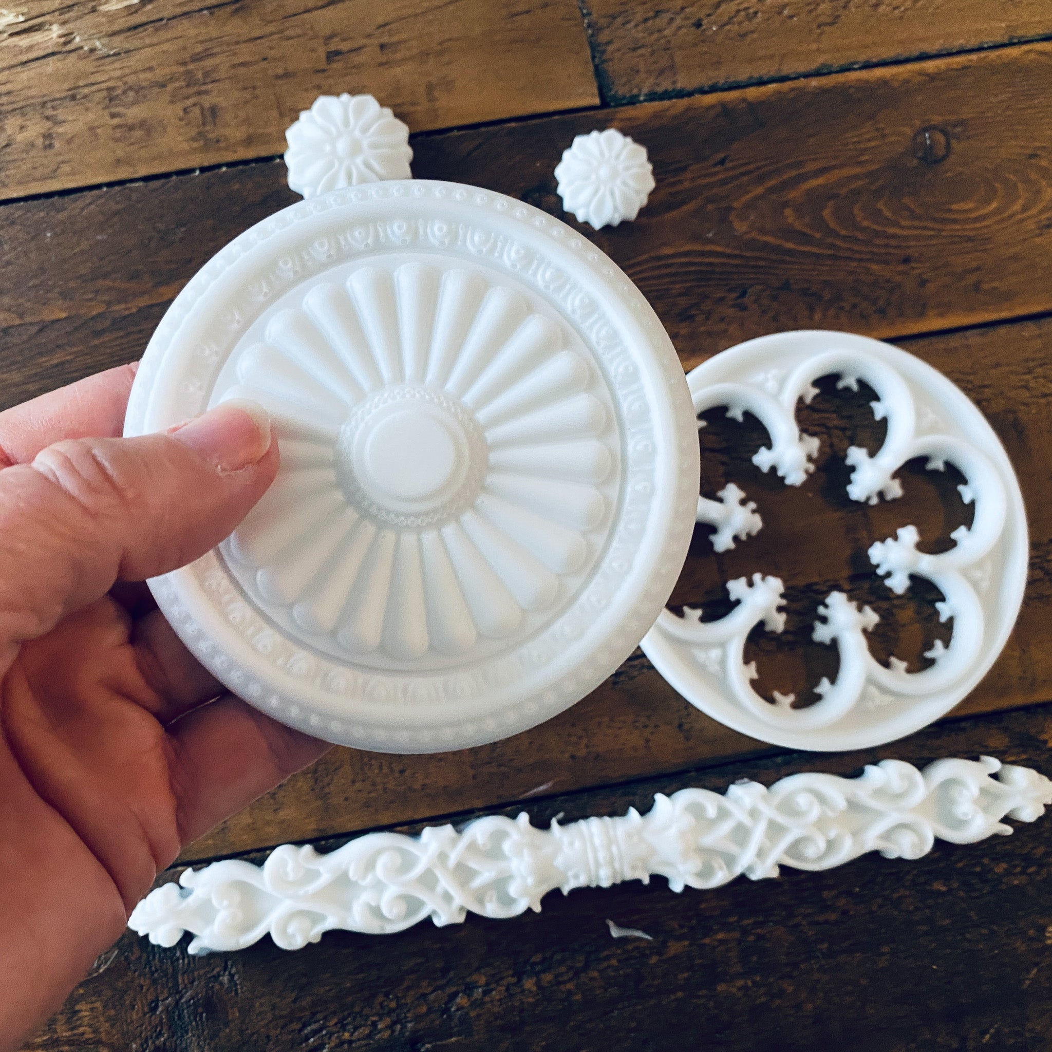 White resin castings of 2 round ornate accent pieces, 2 small round flowers, and a long ornate rod accent piece are against a wood background. A hand is holding one of the larger castings.