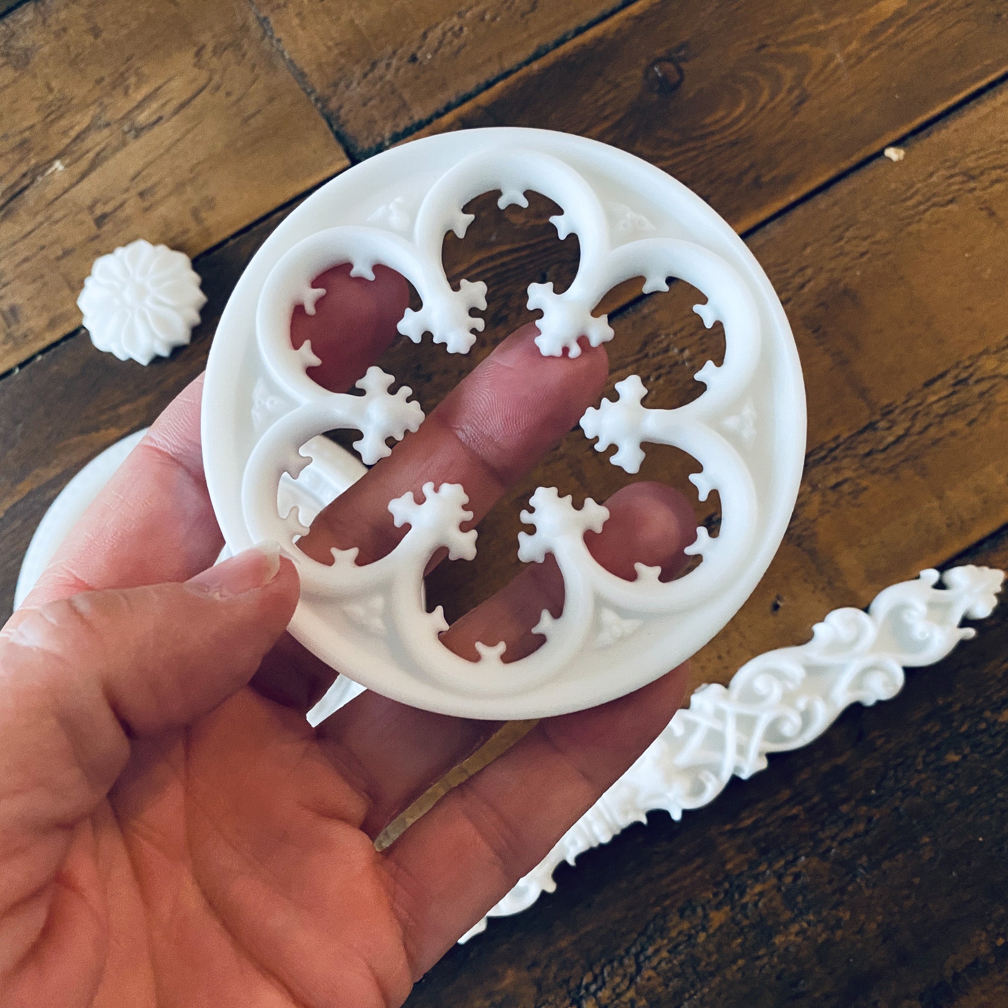 White resin castings of 2 round ornate accent pieces, 2 small round flowers, and a long ornate rod accent piece are against a wood background. A hand is holding one of the larger castings.
