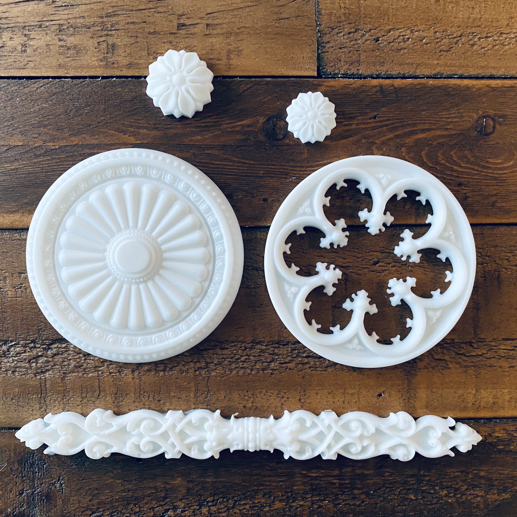 White resin castings of 2 round ornate accent pieces, 2 small round flowers, and a long ornate rod accent piece are against a wood background.