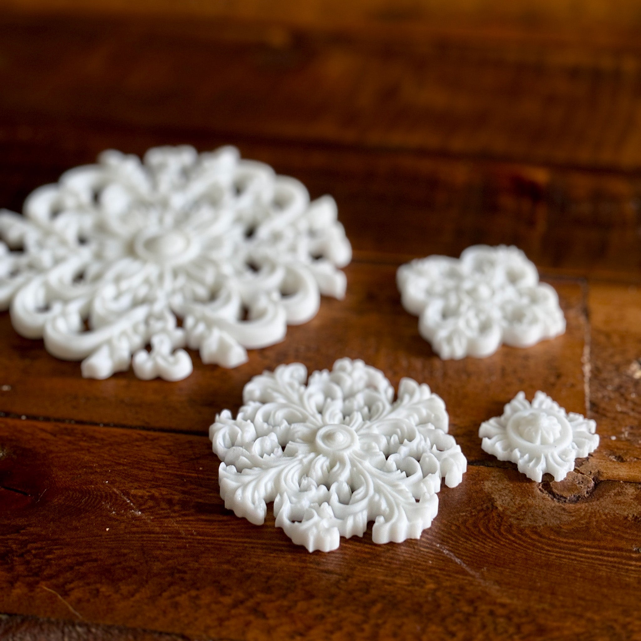 White resin castings of 4 varying size round ornate flourish medallions are against a dark wood background.