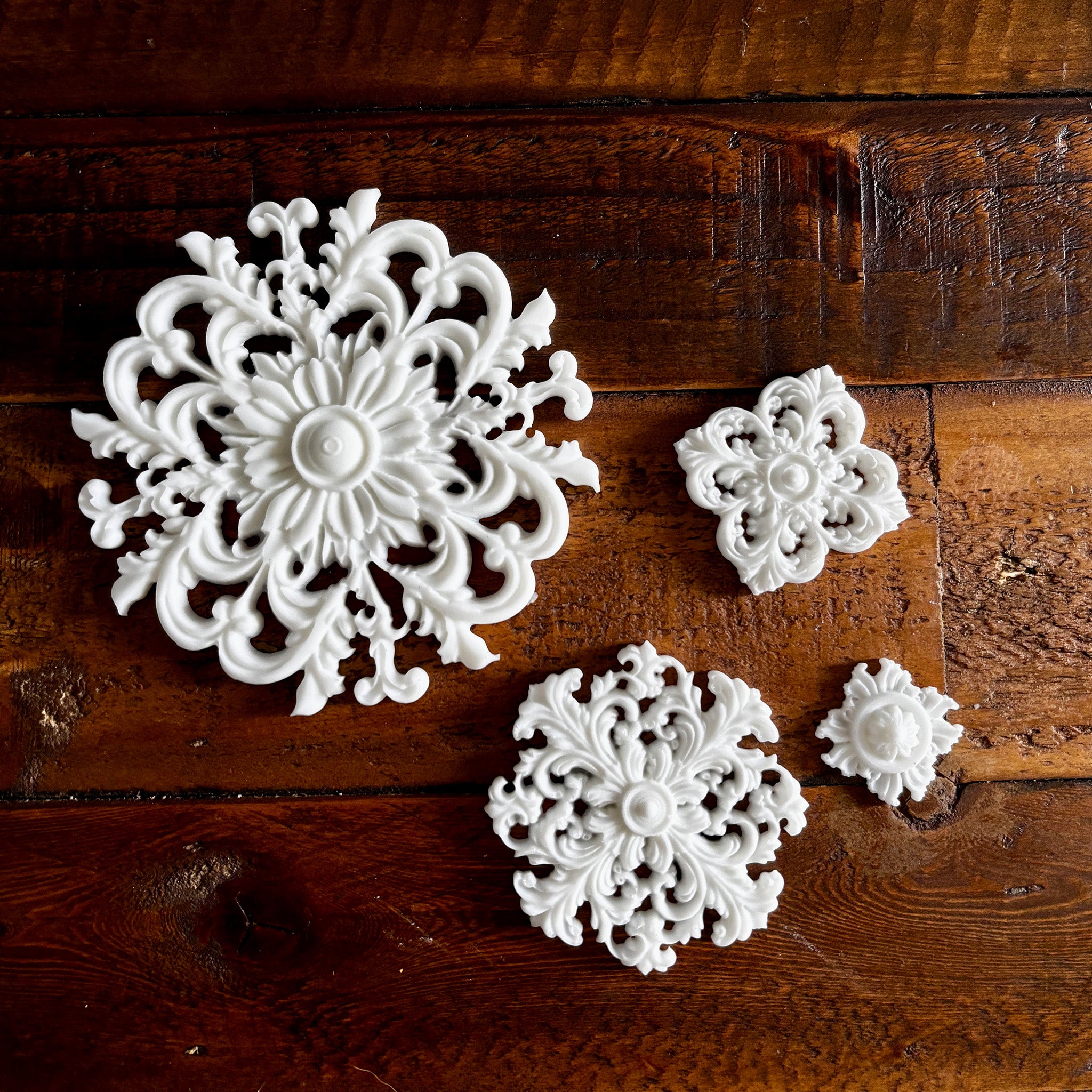 White resin castings of 4 varying size round ornate flourish medallions are against a dark wood background.