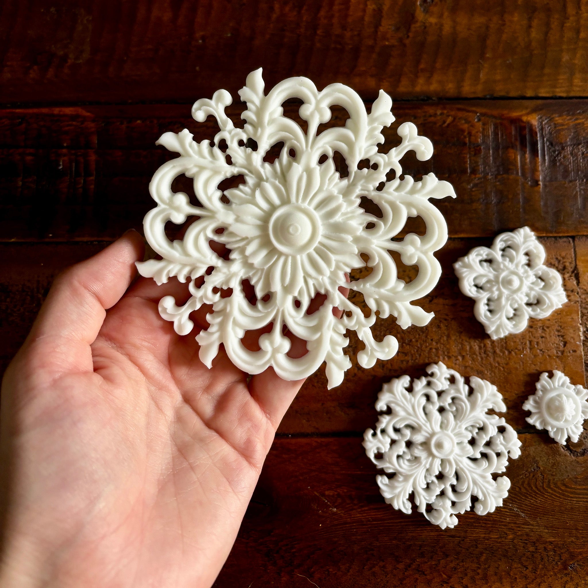 White resin castings of 4 varying size round ornate flourish medallions are against a dark wood background. A hand is shown holding the largest casting.