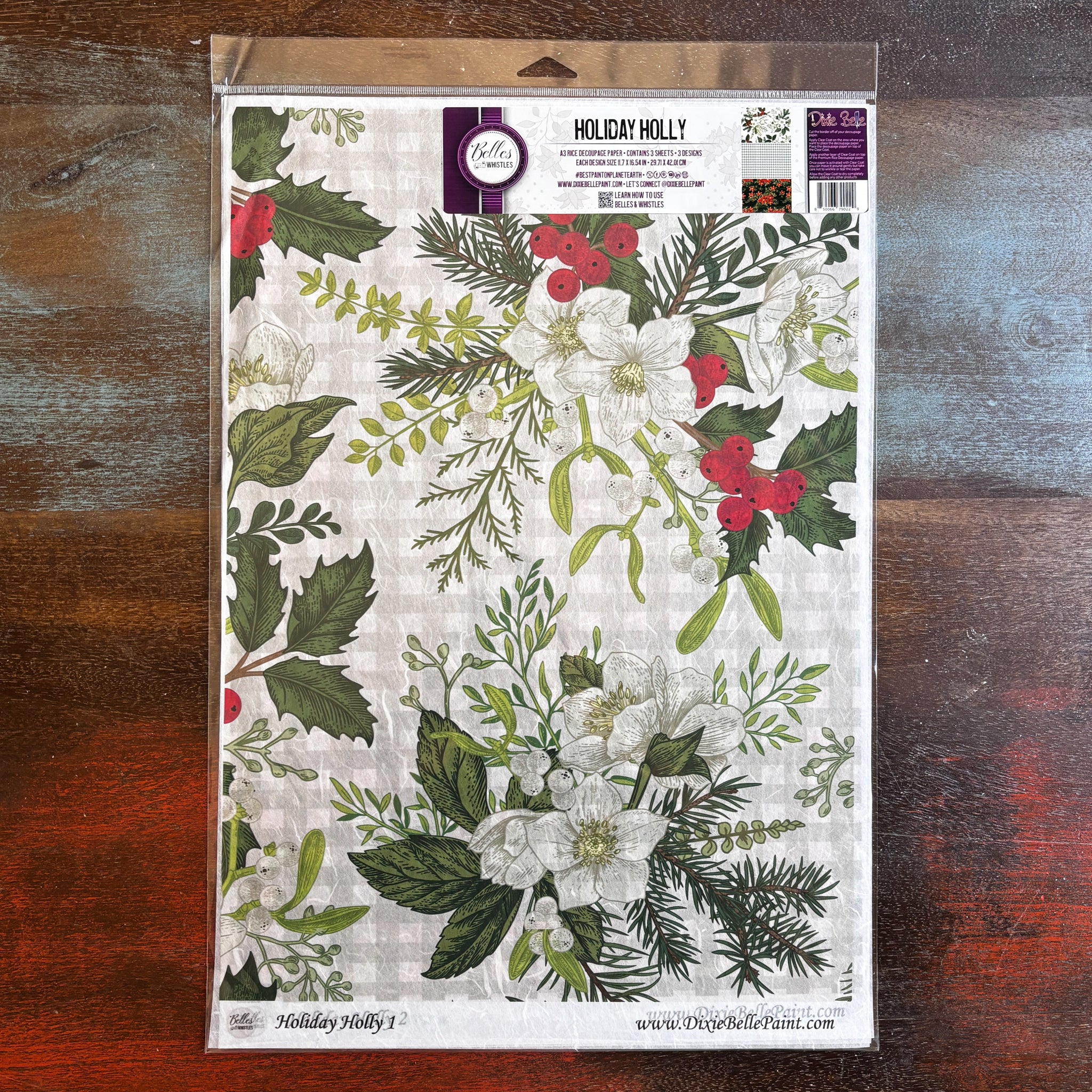 A package of Belles & Whistles' Holiday Holly A3 rice paper is against a dark wood background.