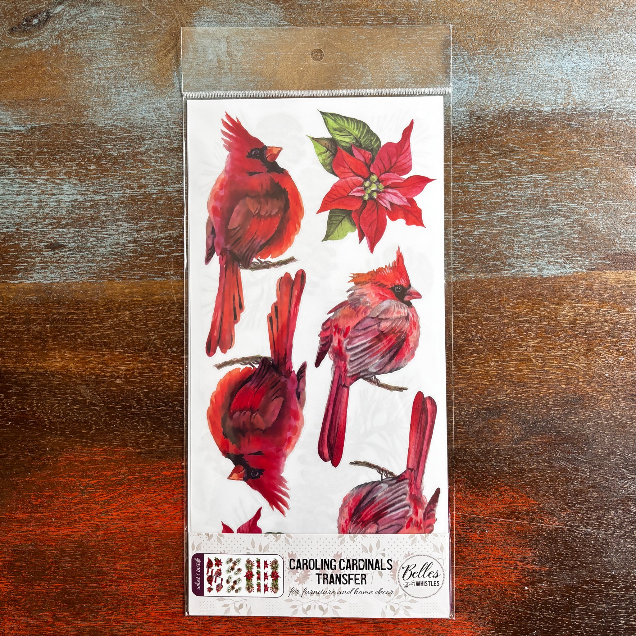 A package of Belles & Whistles' Caroling Cardinals rub-on transfer is against a dark wood background.