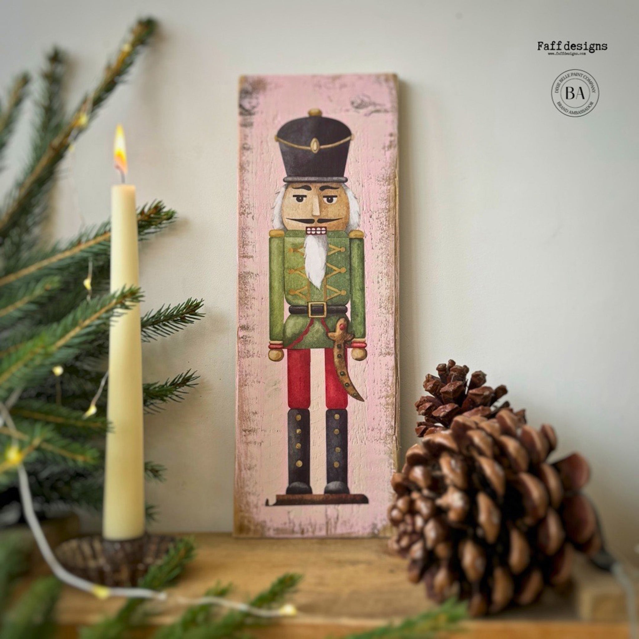 A tall woodcraft project painted pale pink by Faff Designs features a nutcracker in a black hat and green jacket from Belles & Whistles' Nutcracker small rub-on transfer on it.