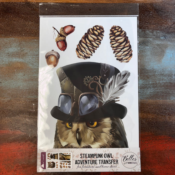 A package of Belles & Whistles' Steampunk Owl Adventures rub-on transfer is against a dark wood background.